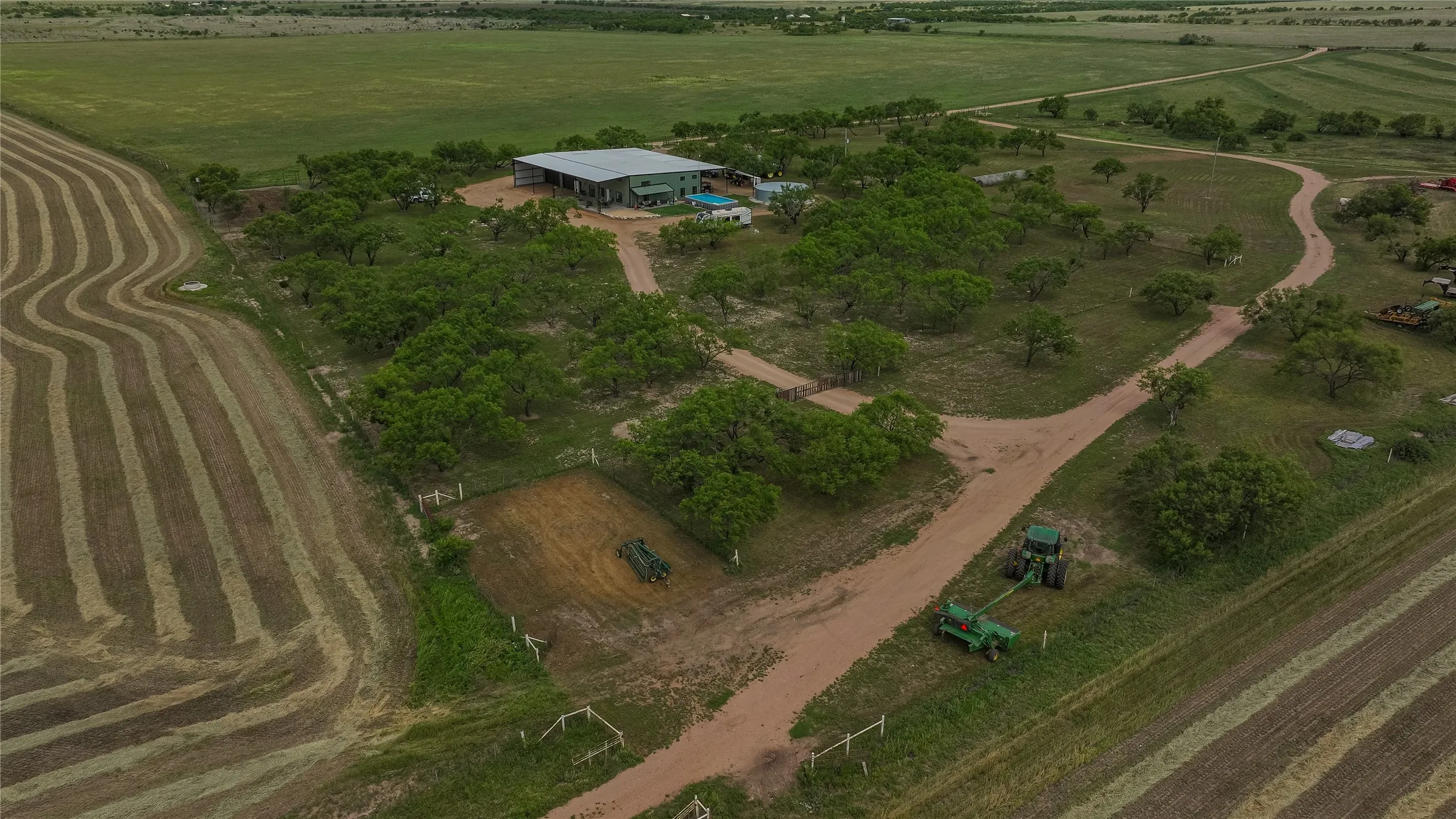Aerial of the homestead with a tractor and hay rake at work. Fenced paddocks, mature mesquite shade and hay fields make this a turnkey working ranch.