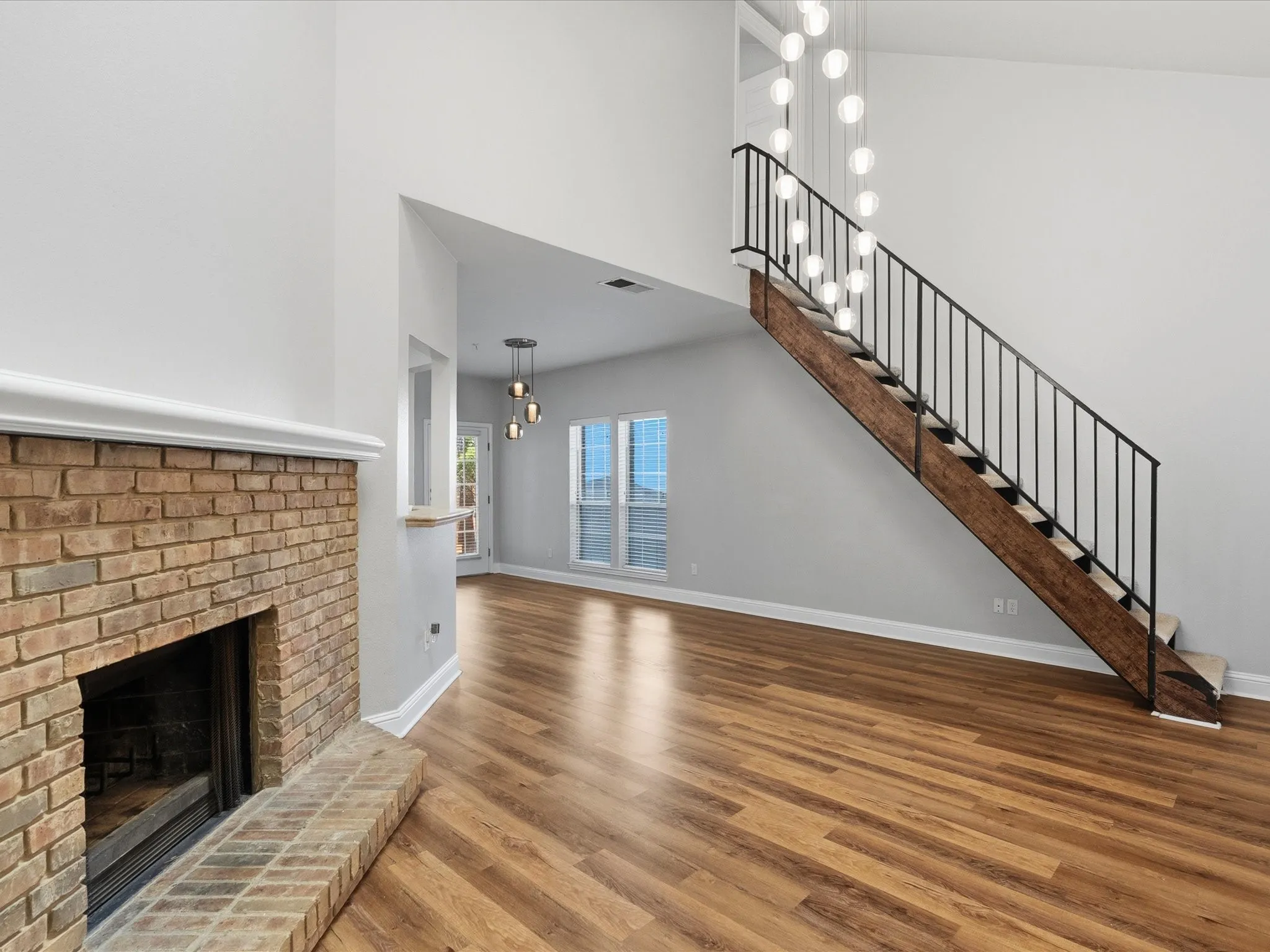 Expansive living area featuring a brick fireplace with a white mantel, wood-finish flooring, and a contemporary staircase with black metal balusters