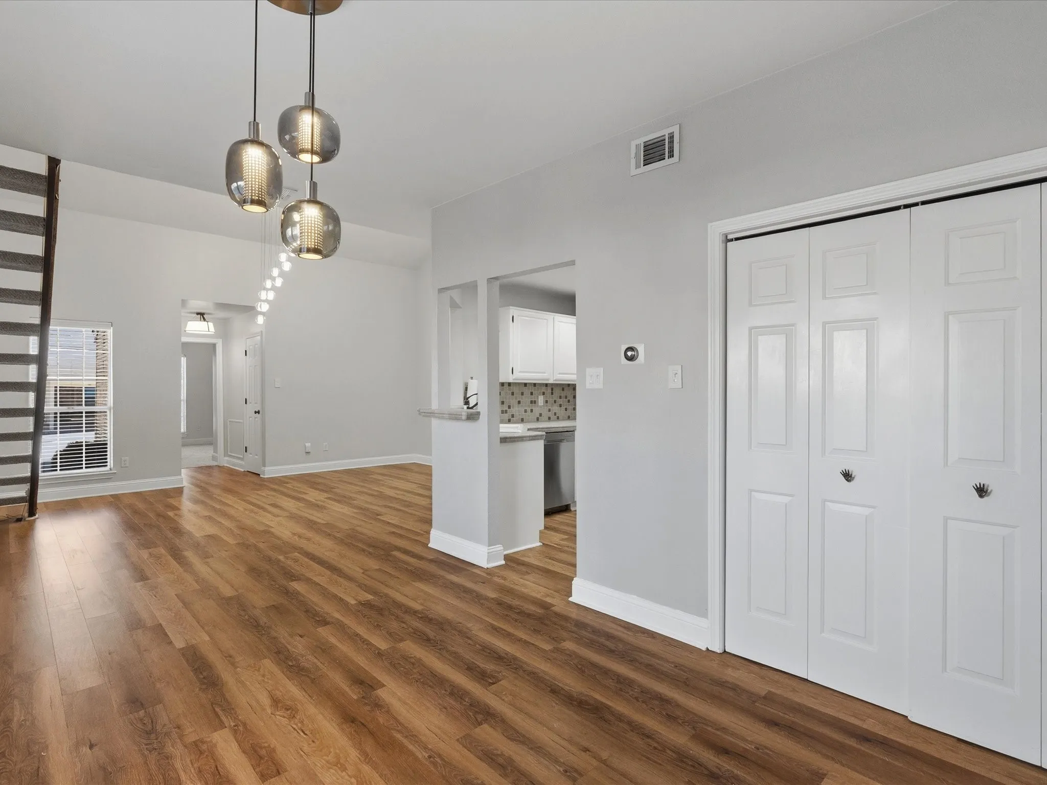 Open-concept living area with wood-finish flooring, a modern three-light pendant fixture, white baseboards, and bi-fold closet doors
