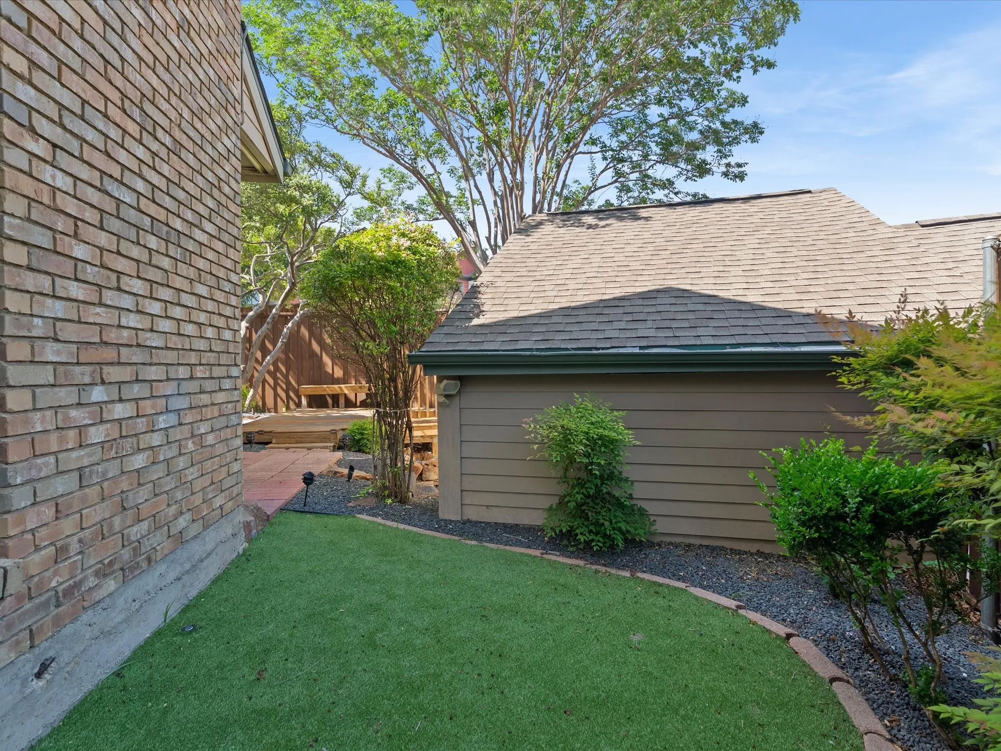Side yard featuring artificial turf, a brick exterior wall, mature landscaping, and a multi-level wood deck
