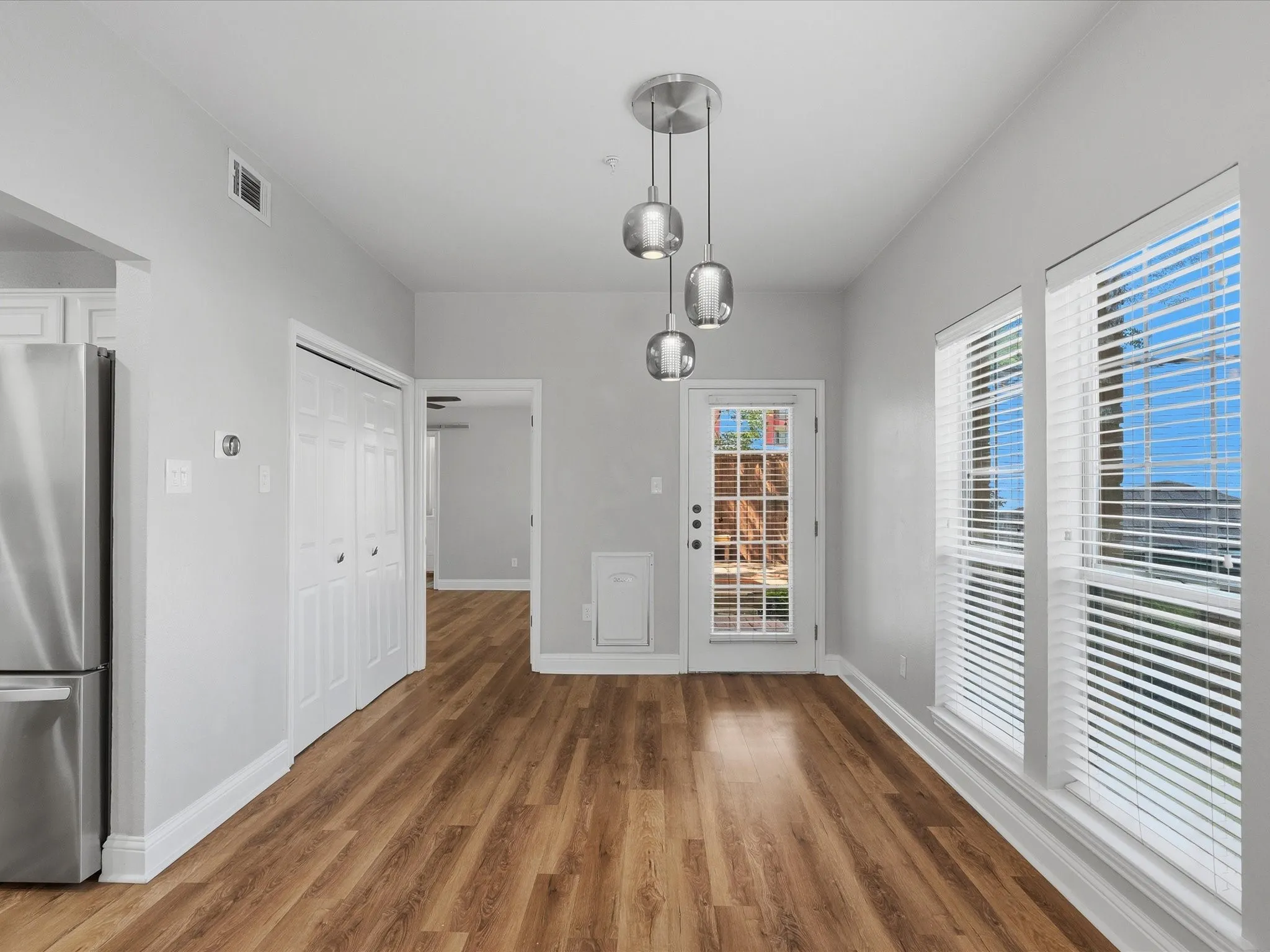 Spacious open plan dining area featuring wood-finish flooring, two large windows with blinds, a modern three-light pendant fixture, a glass-paned exterior door, and a built-in stainless steel​​‌​​​​‌​​‌‌​‌‌​​​‌‌​‌​‌​‌​​​‌​​ refrigerator