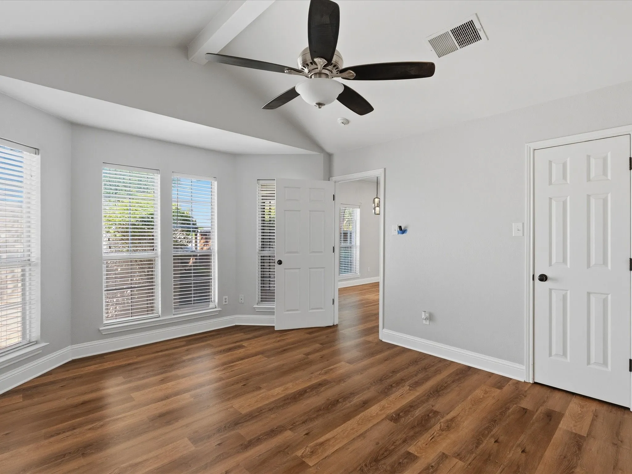Spacious master bedroom featuring wood-finish flooring, light gray walls, a vaulted ceiling with exposed beam accents, and a ceiling fan. Walk in closet and master bath