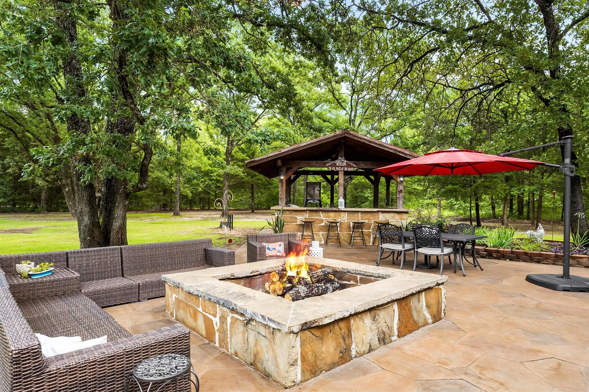 Expansive flagstone patio featuring a built-in fire pit, a covered outdoor bar area, and mature trees providing natural shade