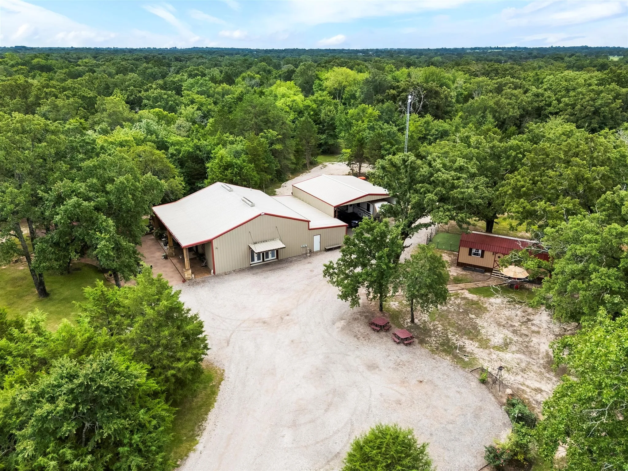 Expansive property featuring a main building with a metal roof and covered porch, a secondary outbuilding, a separate small structure with a red roof, and a large gravel parking area, all surrounded by abundant mature trees