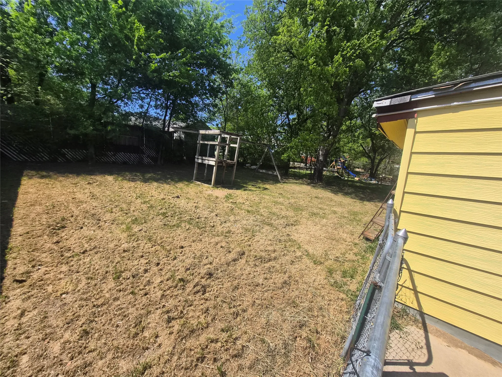 Spacious backyard with mature perimeter trees providing shade, featuring a chain-link fence, and yellow exterior siding