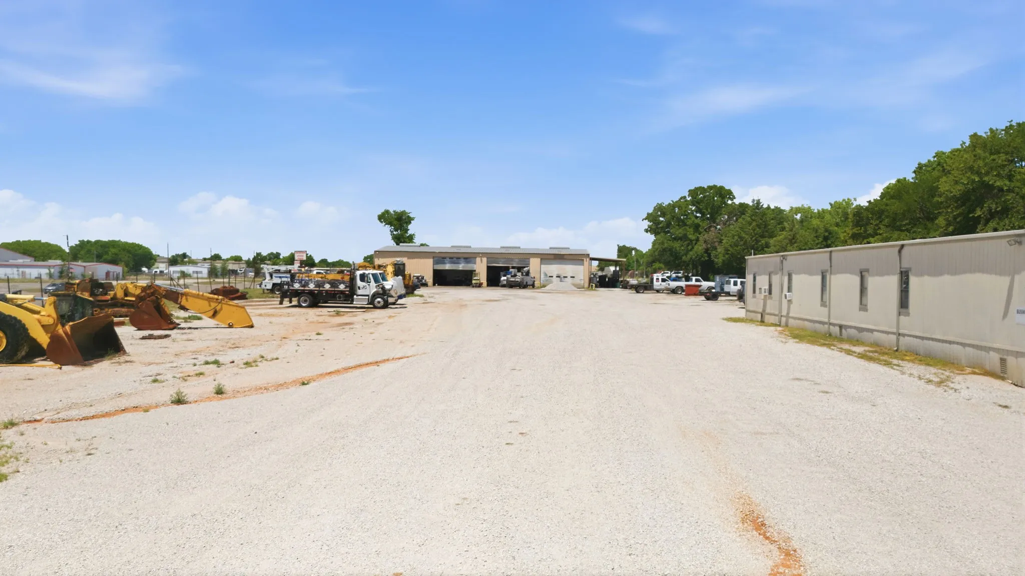 Expansive gravel lot featuring a light-colored commercial building with multiple bays, a secondary portable-style building with multiple windows, and mature trees outlining the property