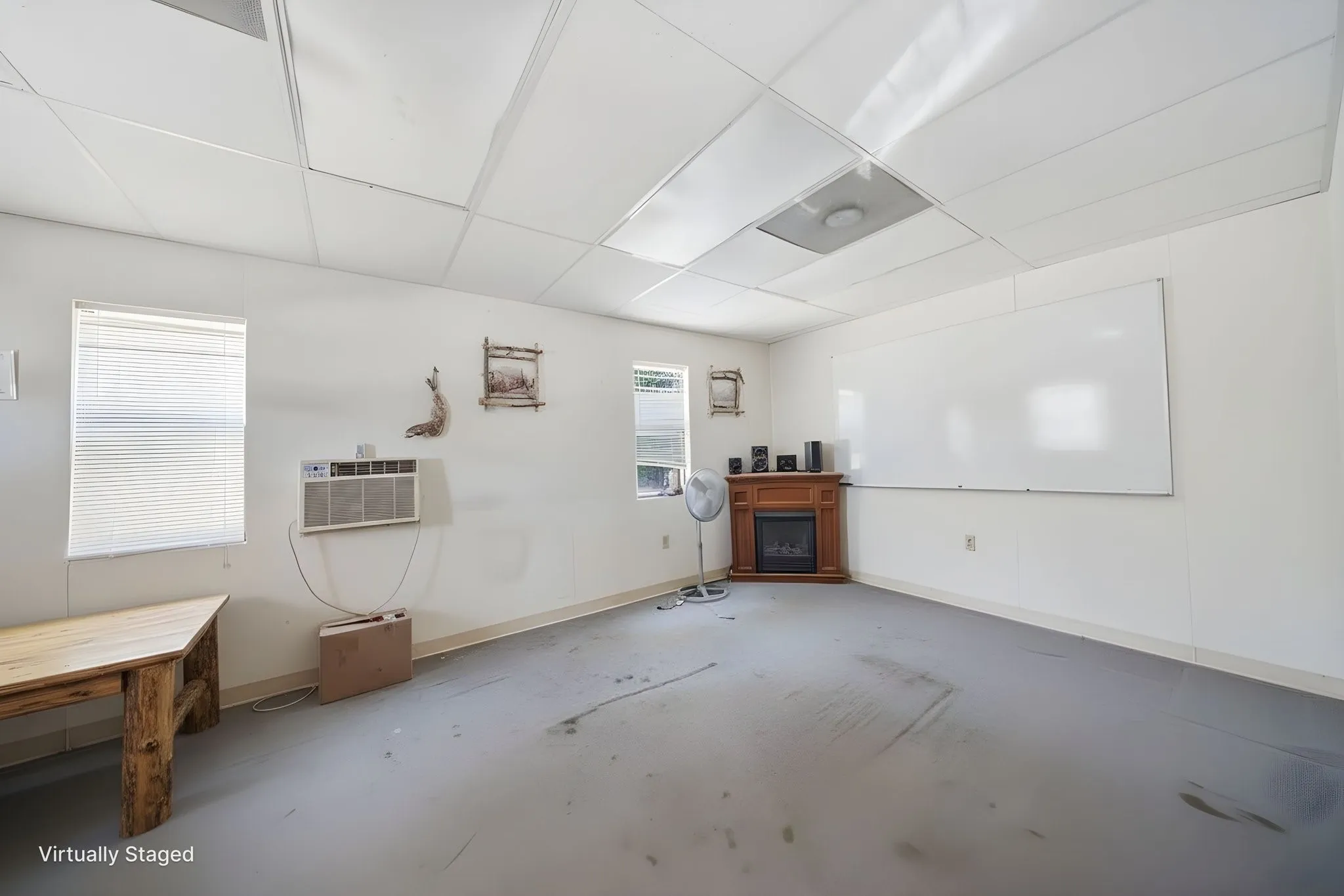 Spacious room featuring a corner-mounted electric fireplace with wood surround, multiple windows with blinds, a large white board, and a white drop ceiling with integrated lighting panels