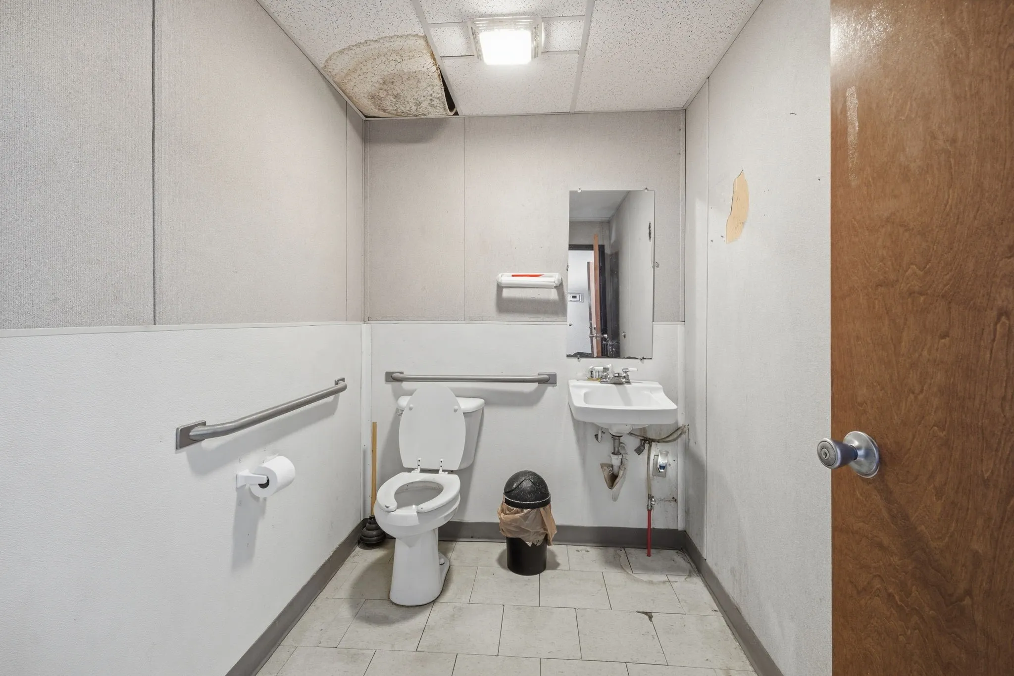 Restroom featuring white square floor tiles, a wall-mounted toilet, a wall-mounted sink, a mirror, and a wood-finish door