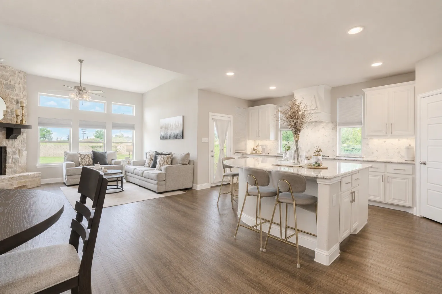Open-concept living area featuring wood-finish flooring, a stone fireplace with a dark wood mantel, and large windows