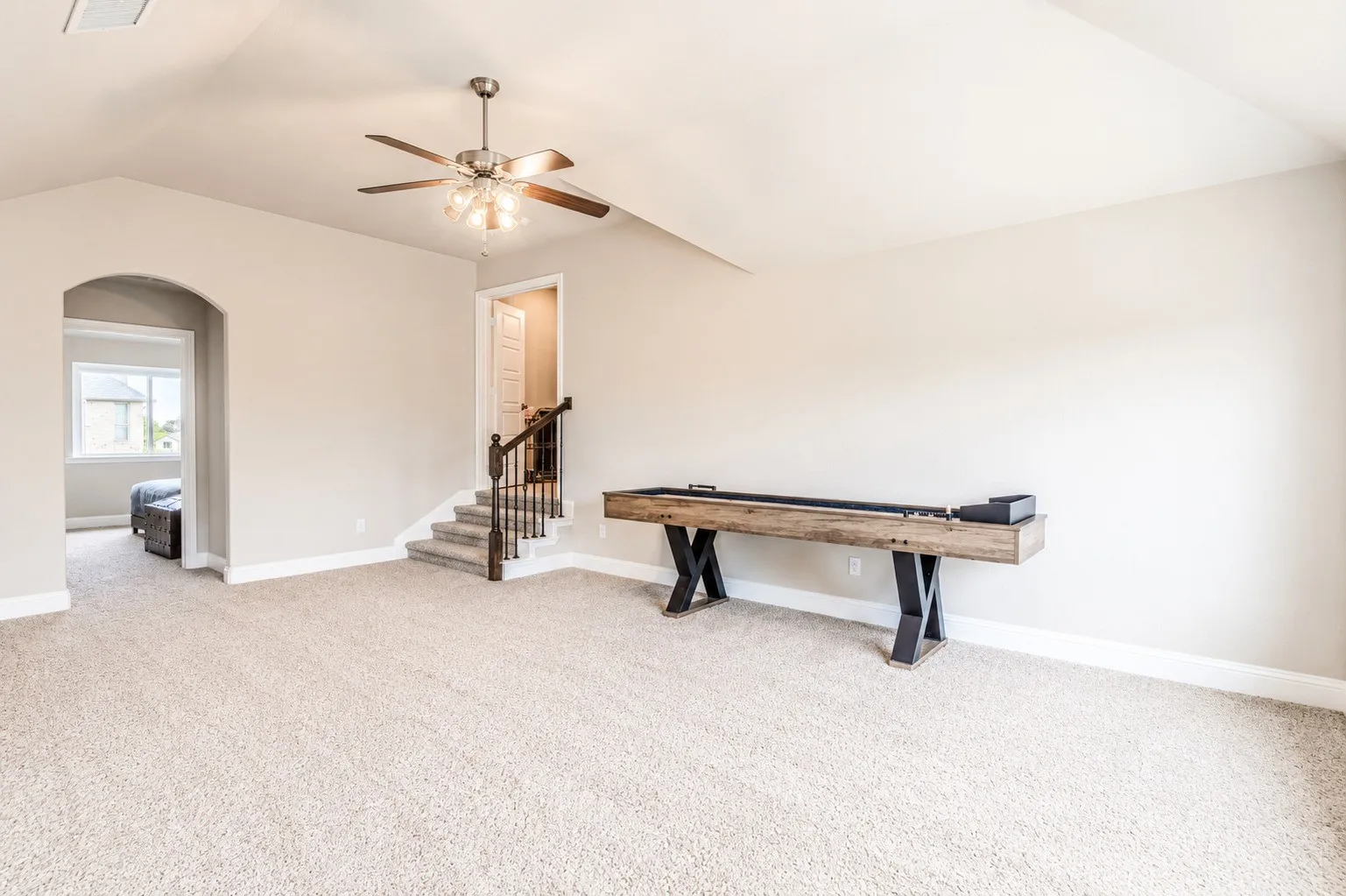 Carpeted bonus room featuring a ceiling fan with light fixture, an arched doorway, and a carpeted staircase with wood handrail and black balusters