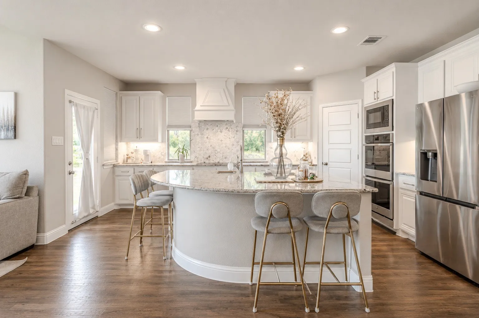 Spacious kitchen featuring a large curved island with a light-toned countertop, wood-finish flooring, white cabinetry, and stainless steel appliances