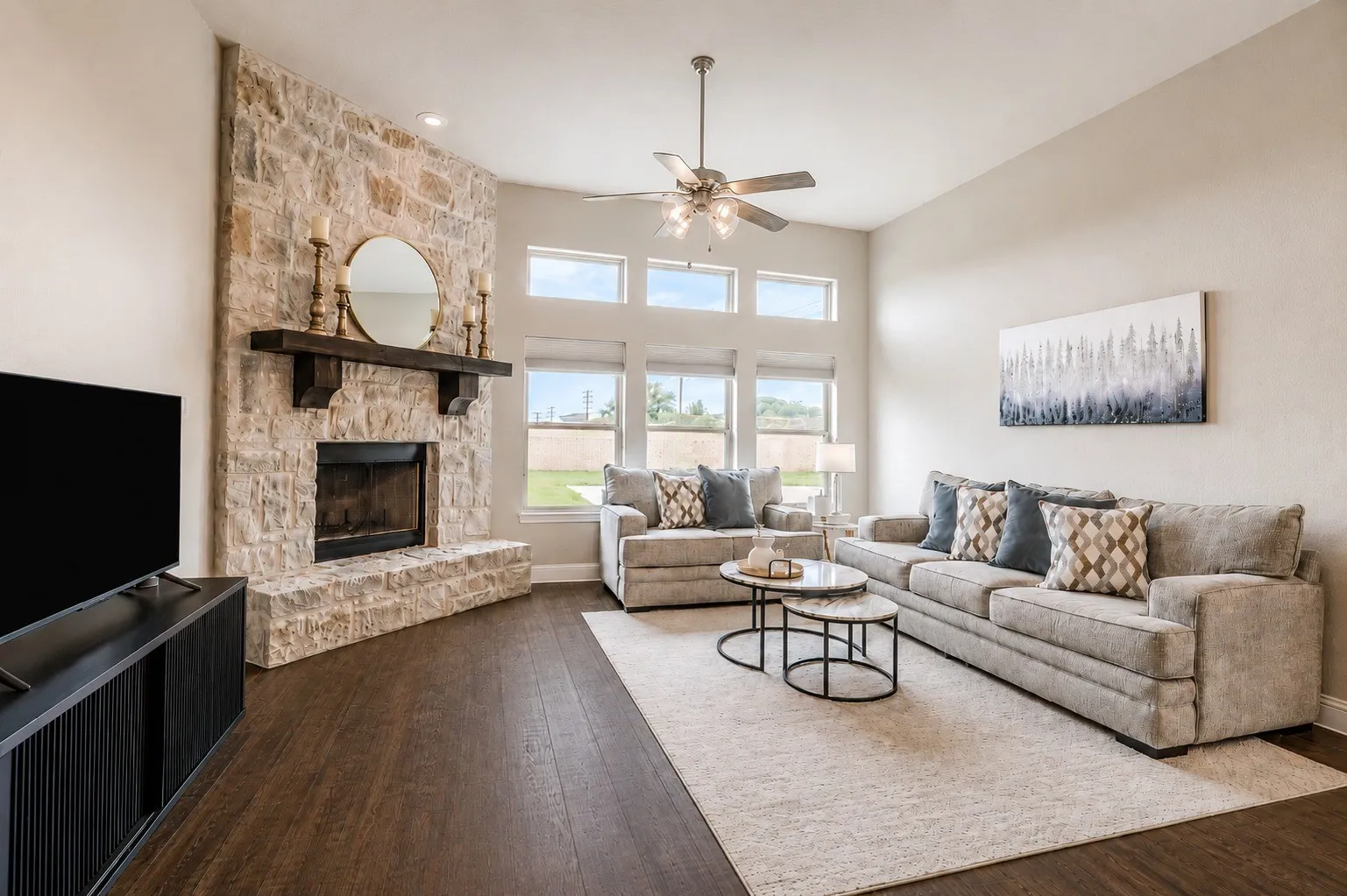 Living space featuring a stone-clad fireplace with a wood mantel, high ceilings, wood-finish flooring, a ceiling fan with integrated lighting, and multiple windows