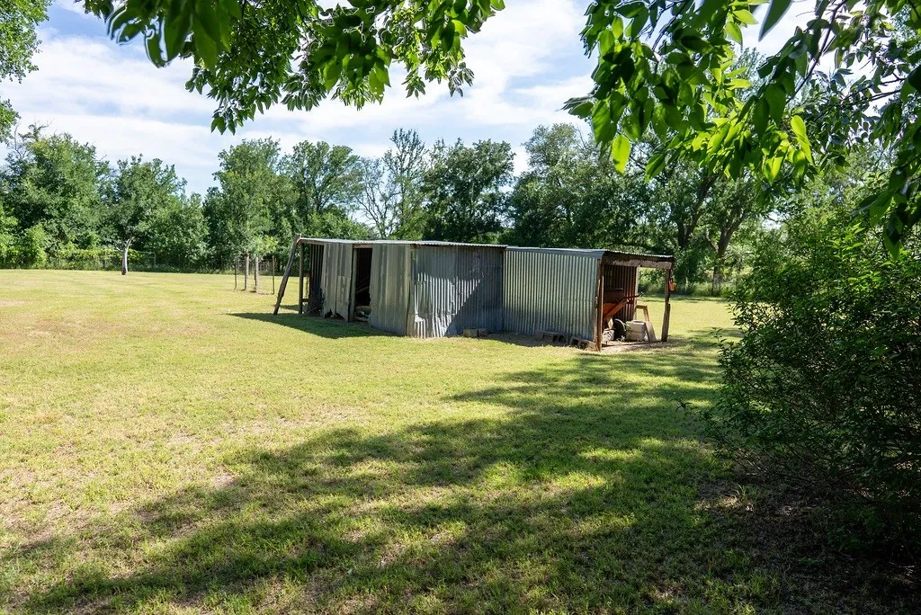 Corrugated metal outbuilding with a partially covered porch area situated on a grass-covered lot with mature trees in the background