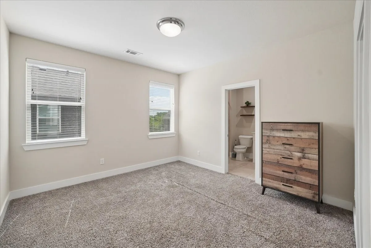 Carpeted bedroom featuring two windows with blinds, a ceiling-mounted light fixture, and a doorway opening to a bathroom with a toilet and floating shelves
