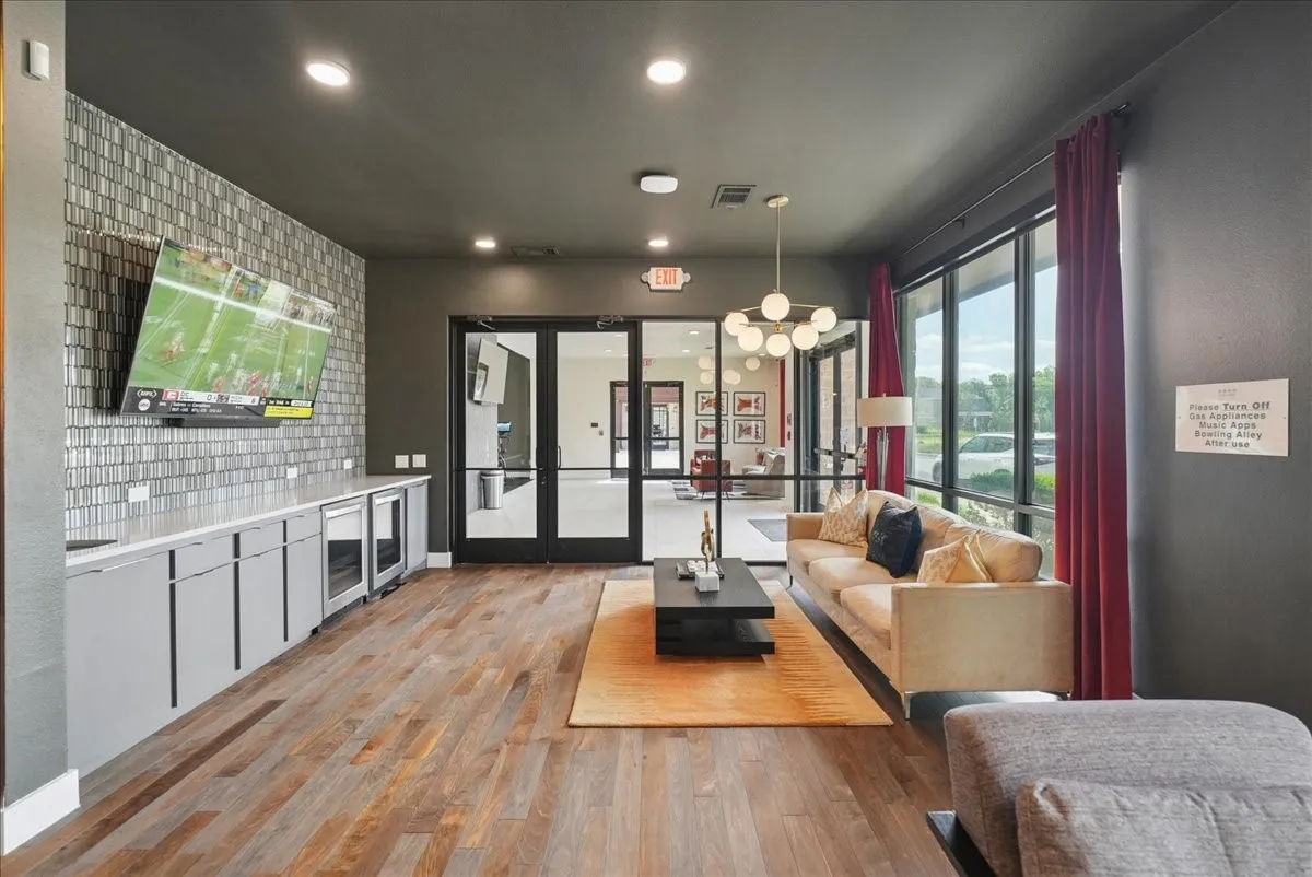 Lounge area featuring wood-finish flooring, a wall-mounted television with a tiled accent wall, built-in cabinetry with integrated beverage coolers, and large windows with full-length drapes