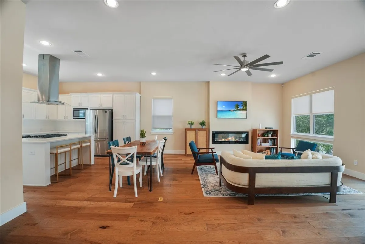 Open-concept living space featuring wood-finish flooring throughout, a kitchen with white cabinetry and stainless steel appliances, a recessed fireplace, a ceiling fan, and large windows with roller shades