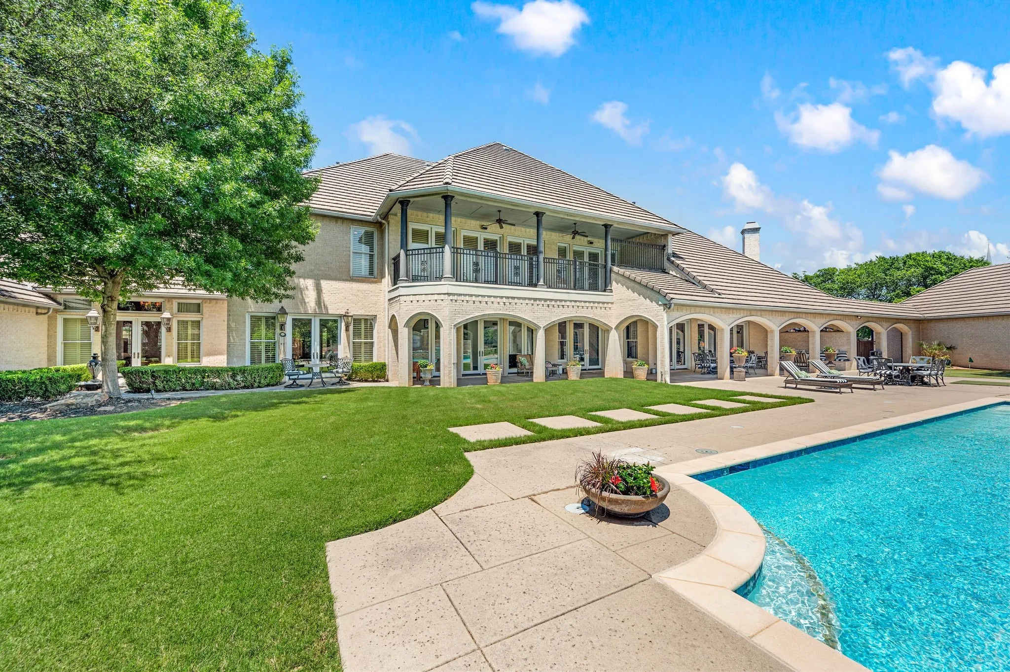Expansive backyard featuring a large swimming pool with integrated spa, a wide paved deck, a manicured lawn, an extended covered patio with multiple arched openings, and a second-story balcony