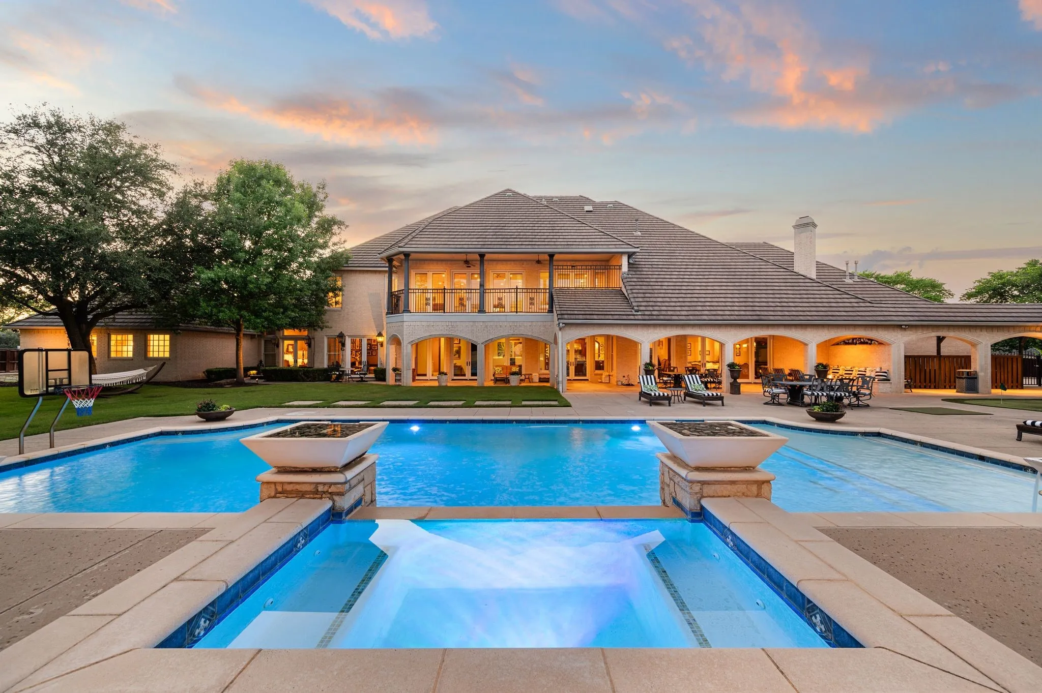 Expansive backyard featuring a large swimming pool with an integrated spa, complemented by a covered patio with arched openings, a grass lawn, and mature trees