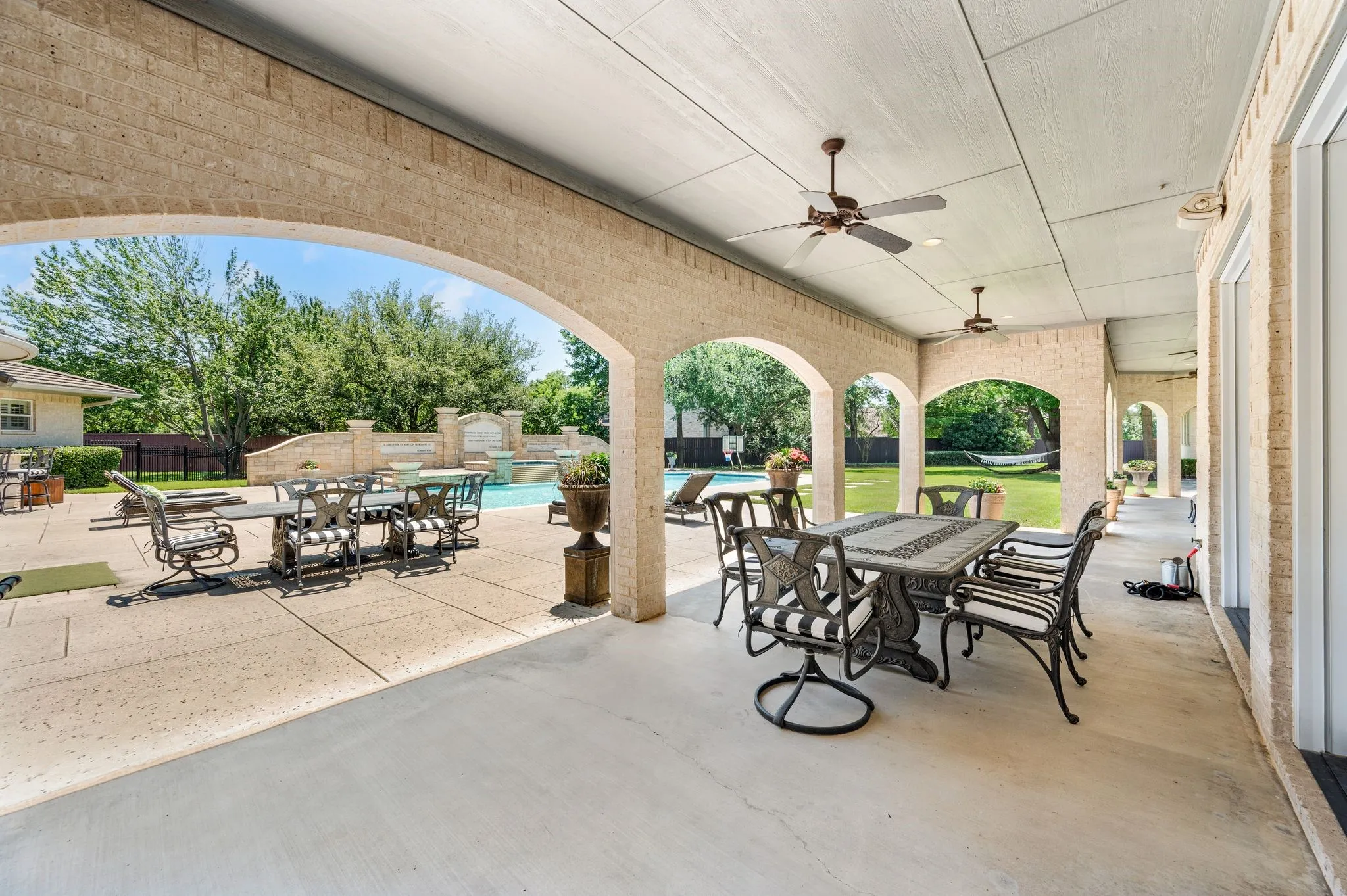 Expansive covered patio featuring brick arches, recessed lighting, and ceiling fans, opening onto a concrete patio with a swimming pool and lush green lawn