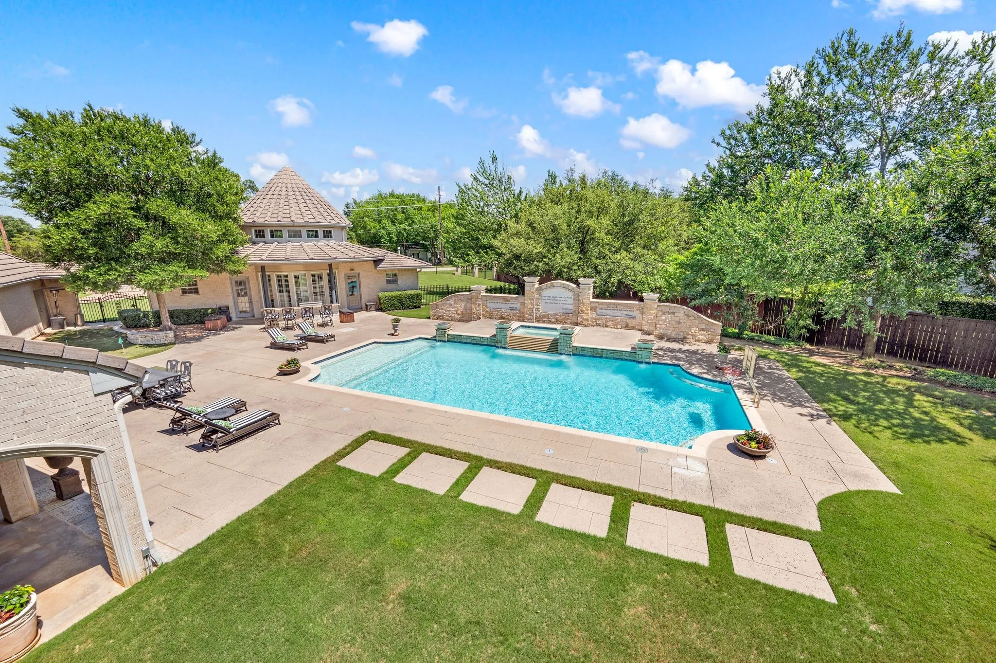 Expansive backyard featuring a concrete patio, an inground swimming pool with integrated spa, a stone retaining wall with water features, a green lawn area with stepping stones, and mature perimeter trees
