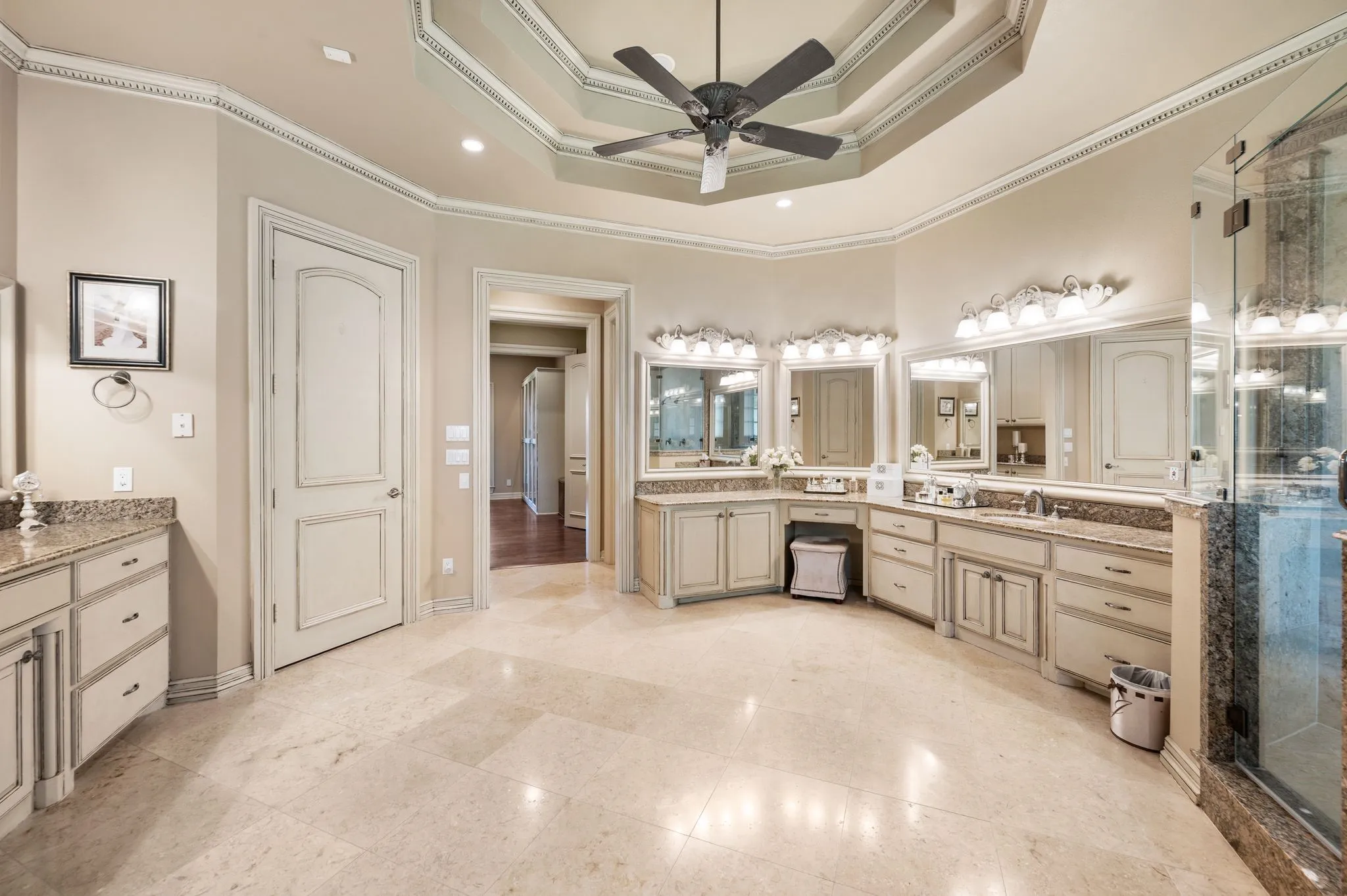 Spacious bathroom featuring light-toned flooring, an ornate tray ceiling with a ceiling fan, and extensive vanity space with light cabinetry and stone countertops