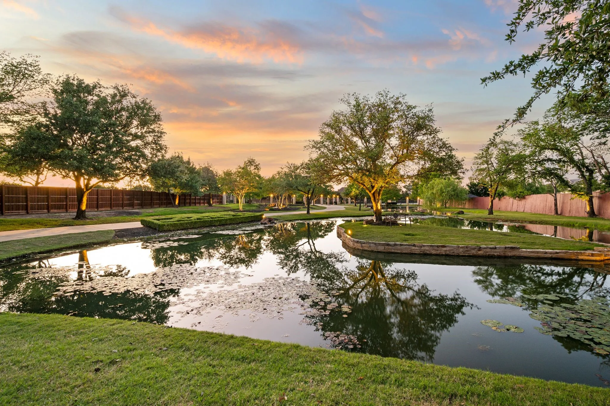 Tranquil water feature with illuminated trees, an island with manicured greenery, and a paved walking path alongside a privacy fence