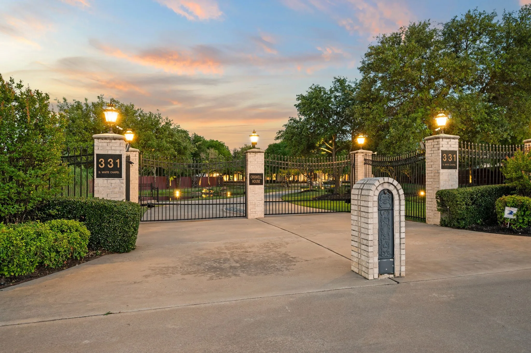 Gated entry featuring stone columns with integrated lighting, wrought iron fencing, and manicured landscaping
