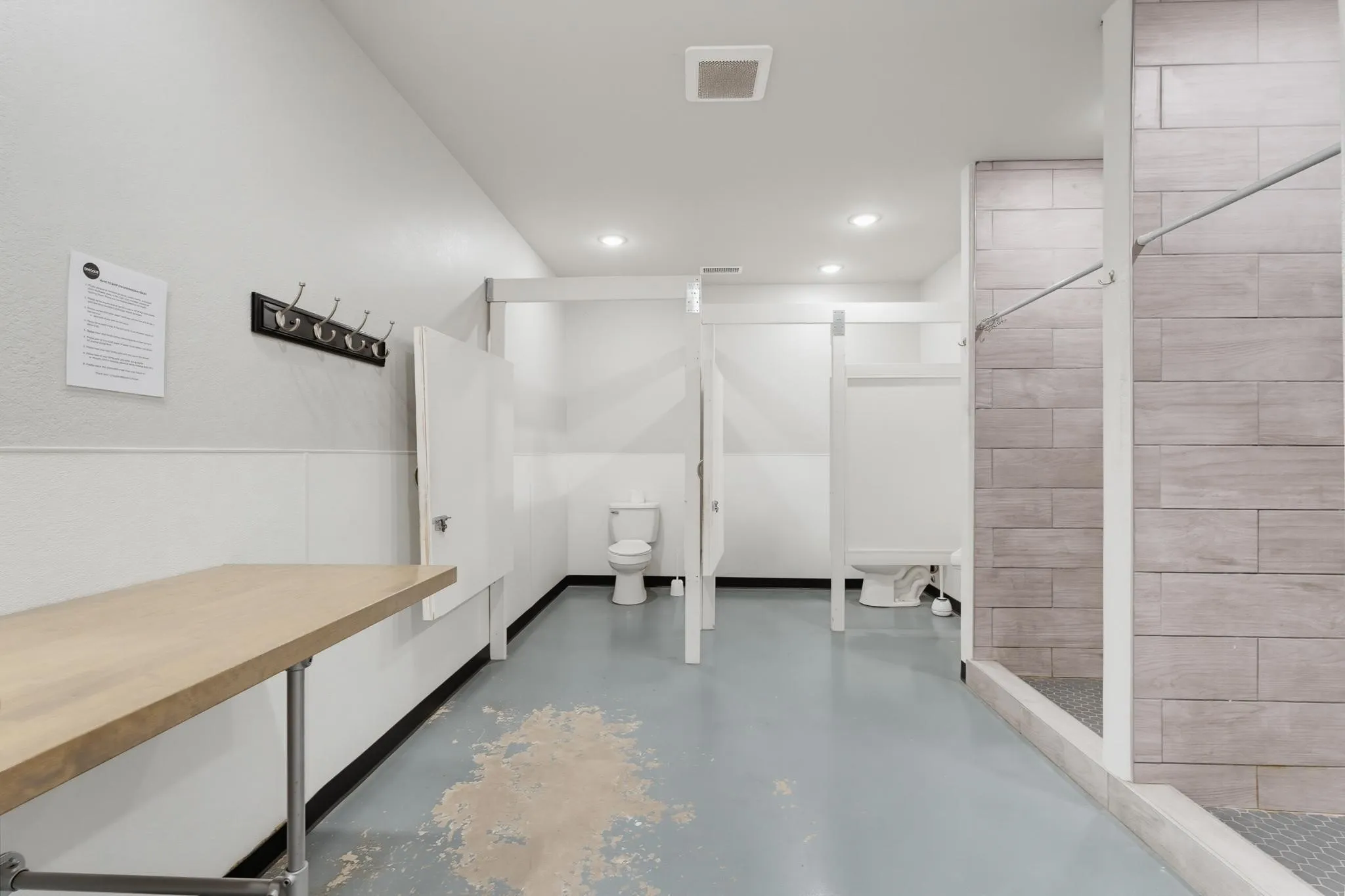 Commercial restroom with individual toilet stalls, a built-in shower, and a light wood-finish counter
