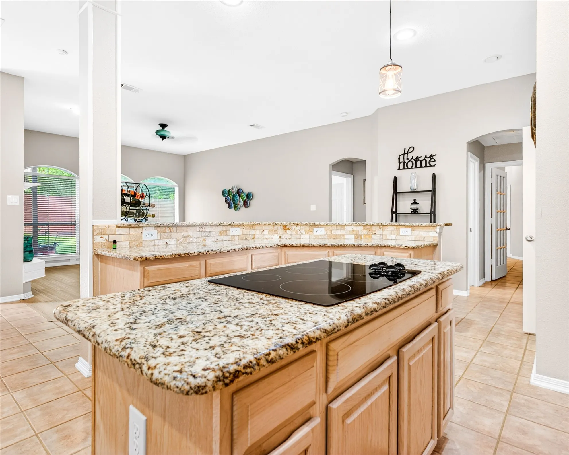 Kitchen island featuring a built-in electric cooktop, granite countertops, light wood-finish cabinetry, and a stone-tiled backsplash