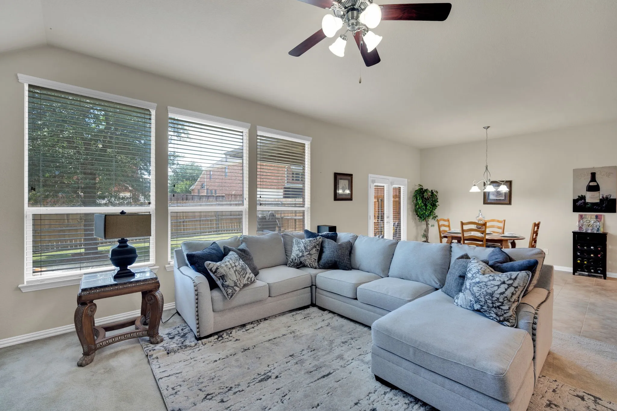 Spacious living area featuring neutral wall tones, multiple large windows with blinds, a ceiling fan with light fixture, and tile flooring extending towards the dining area