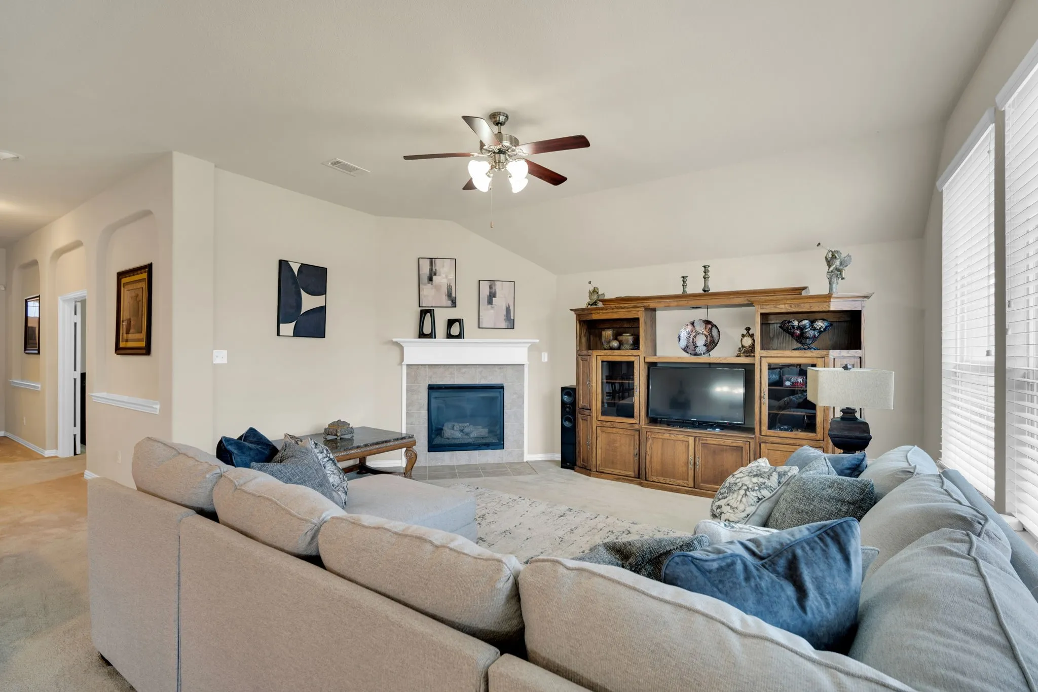 Spacious living area featuring a vaulted ceiling with a ceiling fan, a fireplace with a white mantel and tile surround, and tall windows with blinds