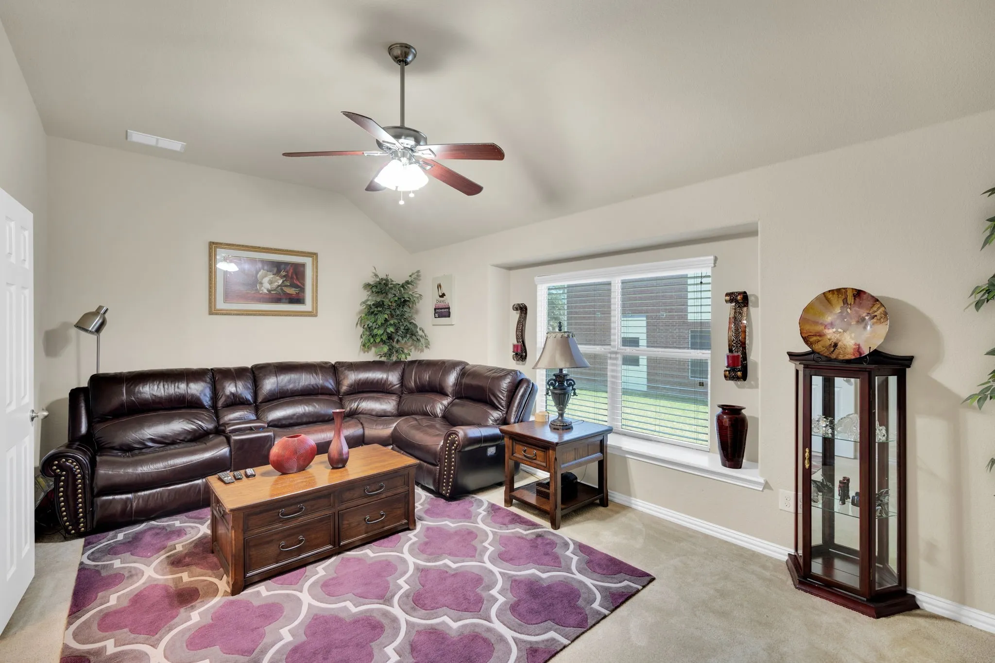 Living area with vaulted ceiling and ceiling fan, featuring a large window, neutral wall color, and carpet flooring