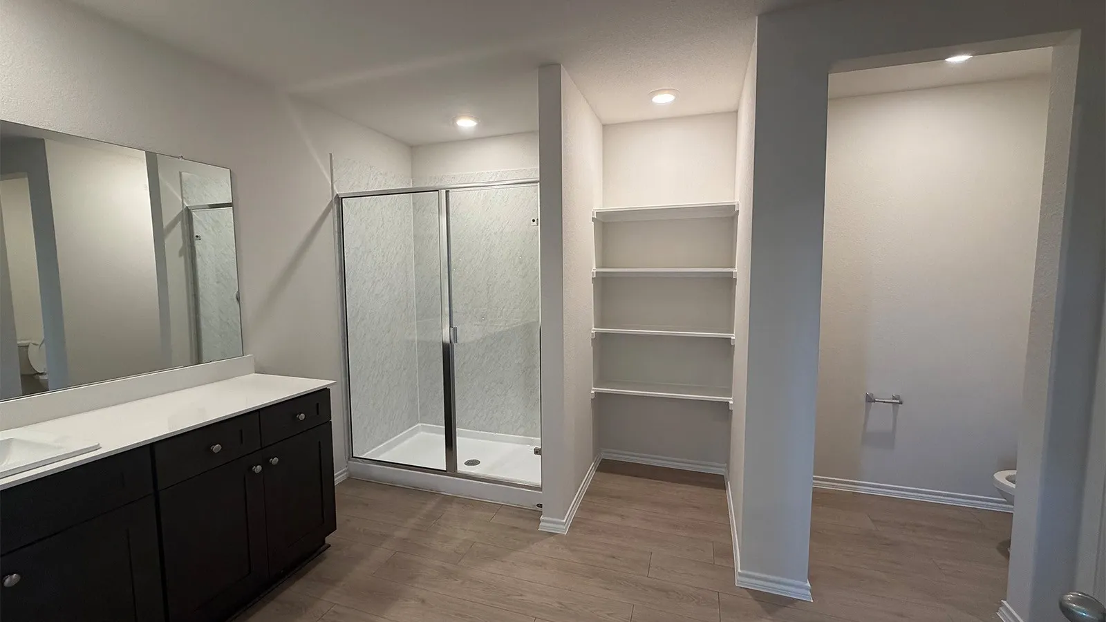 Bathroom featuring a dual vanity with dark wood cabinetry and white countertops, a glass-enclosed shower, built-in shelving, and wood-finish flooring