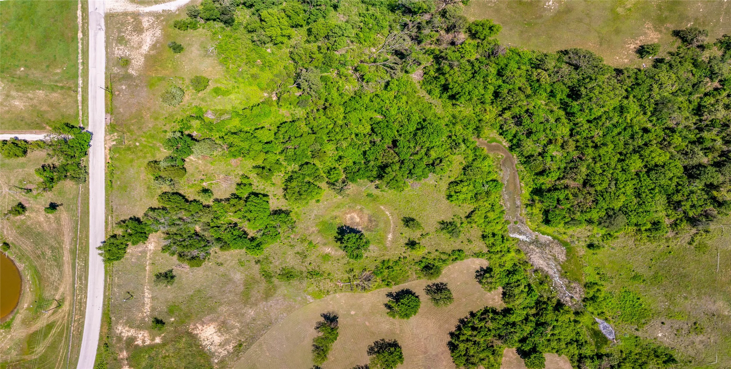 Expansive land parcel featuring a paved road frontage, natural tree coverage, open grassy areas, and a visible water feature