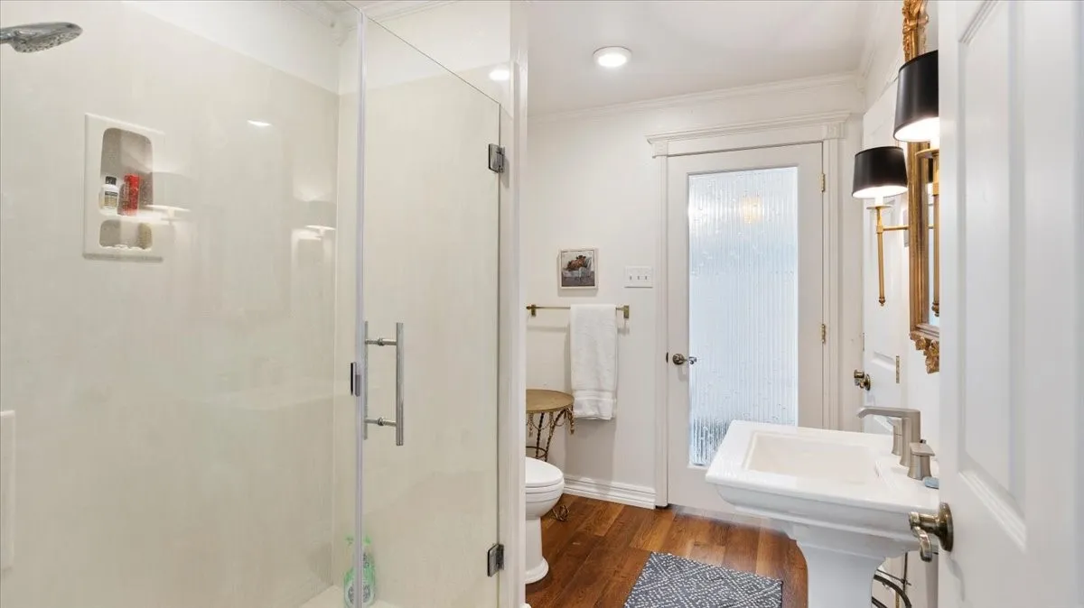 Bathroom featuring a frameless glass shower enclosure, a recessed shower niche, a pedestal sink with a contemporary faucet, wood-finish flooring, and a door with privacy glass