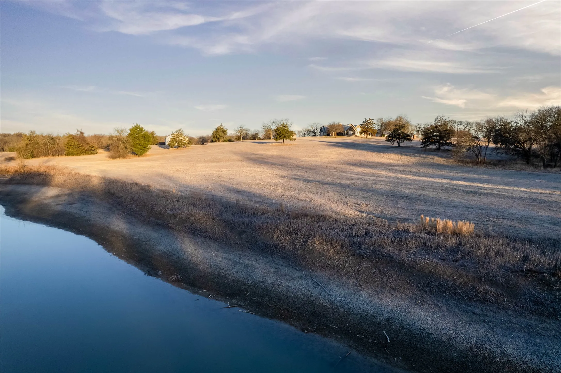 Expansive property featuring a body of water, rolling terrain, scattered trees, and distant structures