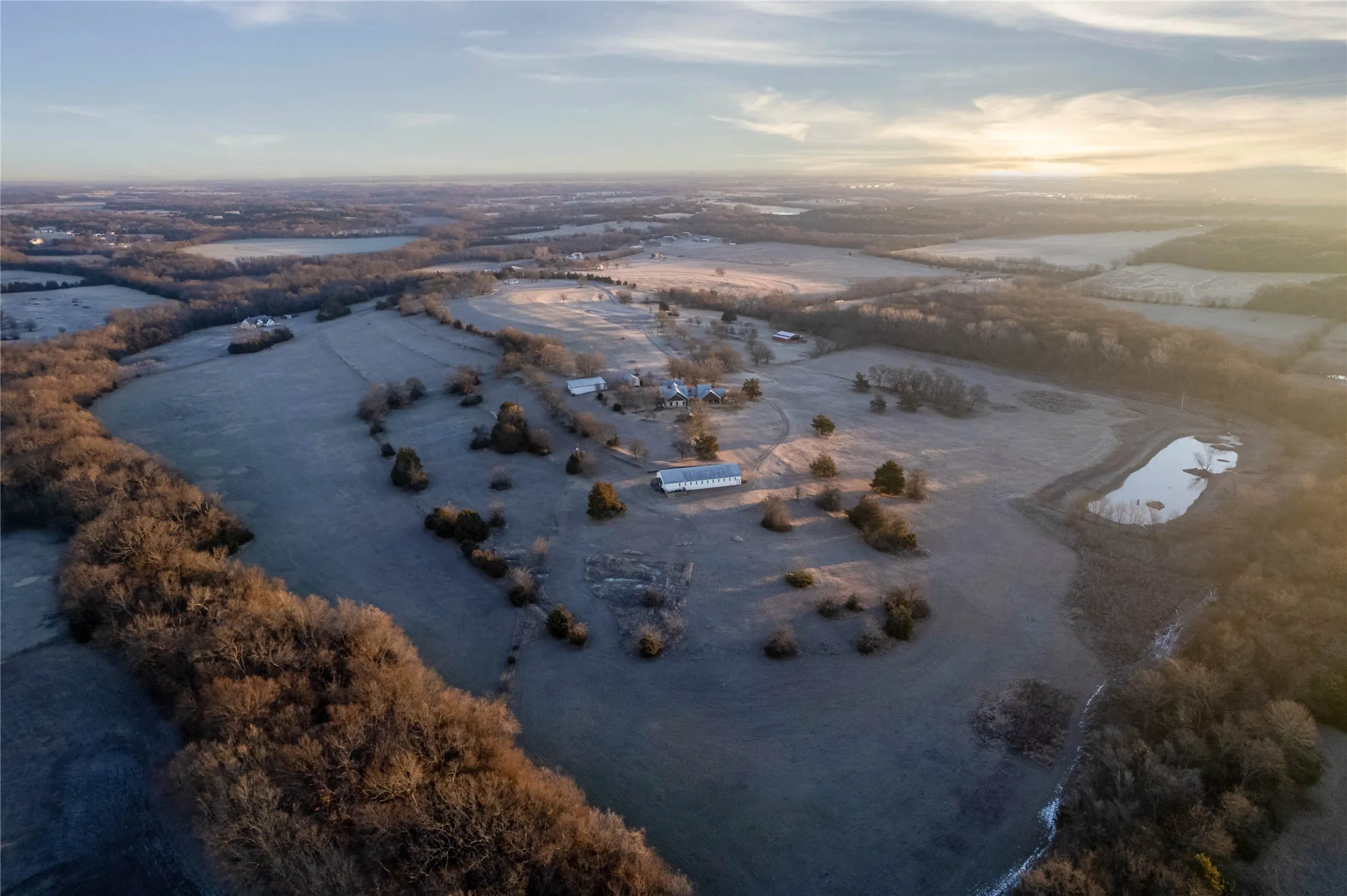 Expansive land featuring a natural pond, scattered mature trees, and multiple outbuildings