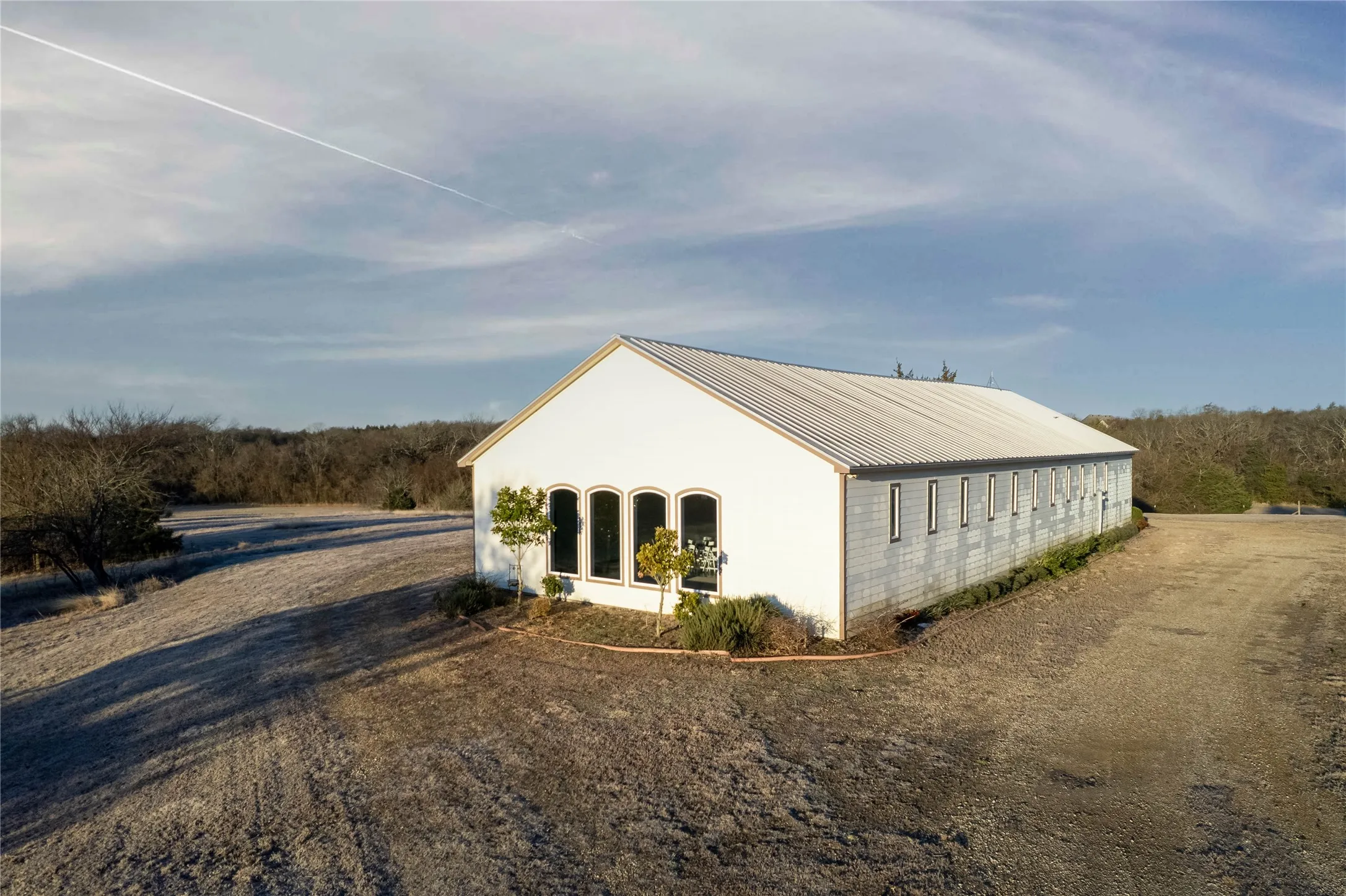 White-sided building with a metal roof, featuring multiple arched windows at the front and a series of rectangular windows along the side