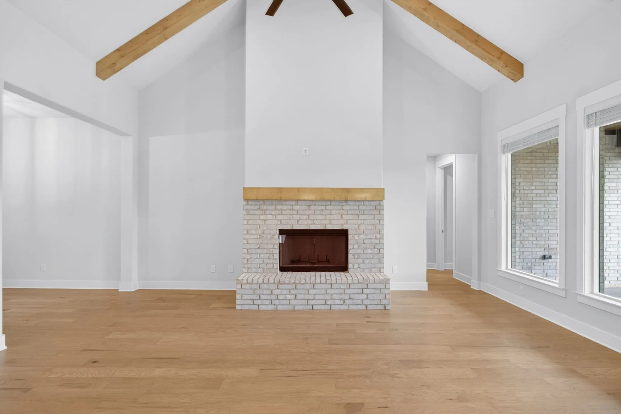 Vaulted ceiling with exposed wood beams, a white brick fireplace with a wood mantle, light wood-finish flooring, and multiple windows