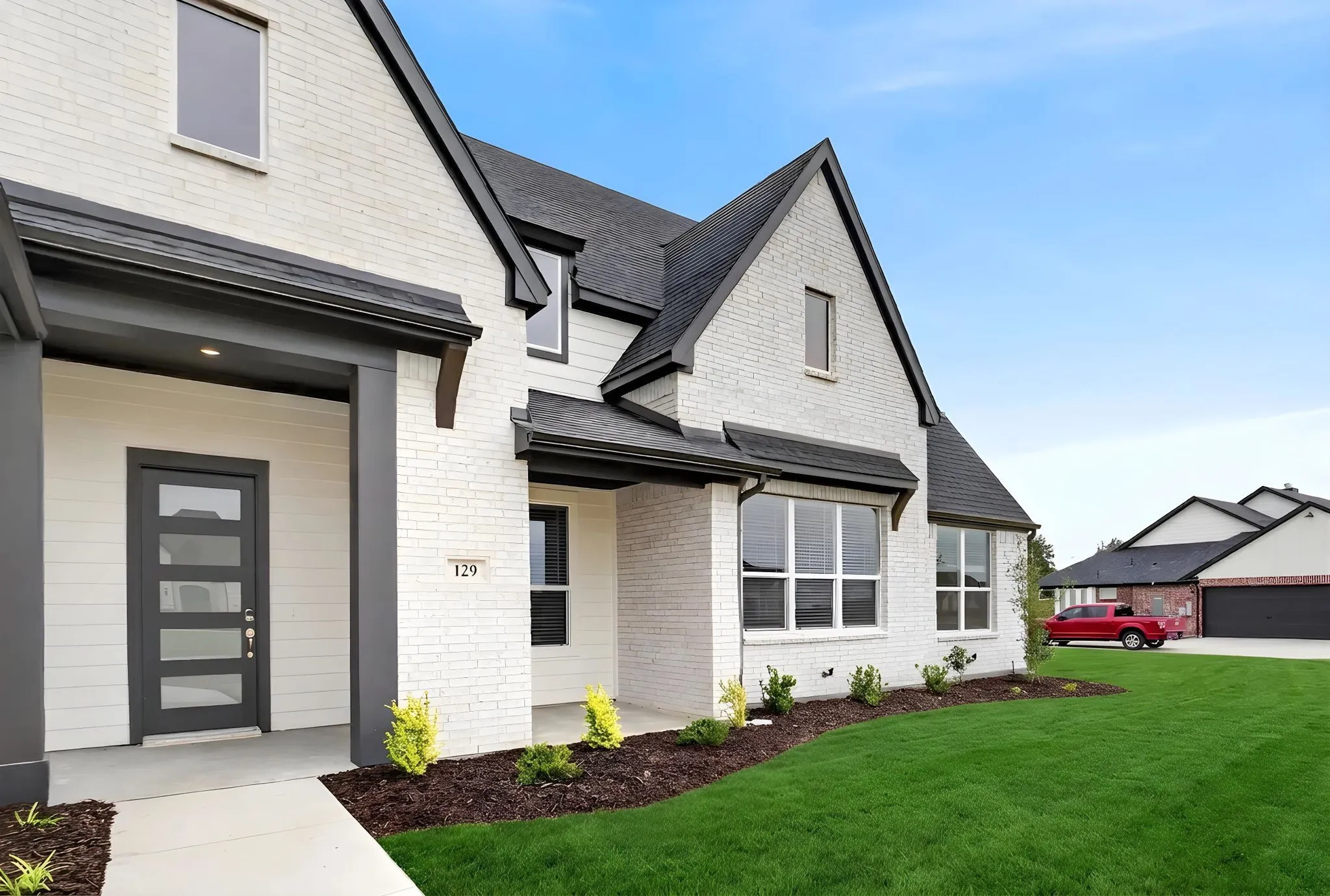 Contemporary exterior featuring white brick and siding, a dark gray entry door with multiple glass panes, black-trimmed windows, a dark shingle roof, and a meticulously manicured lawn