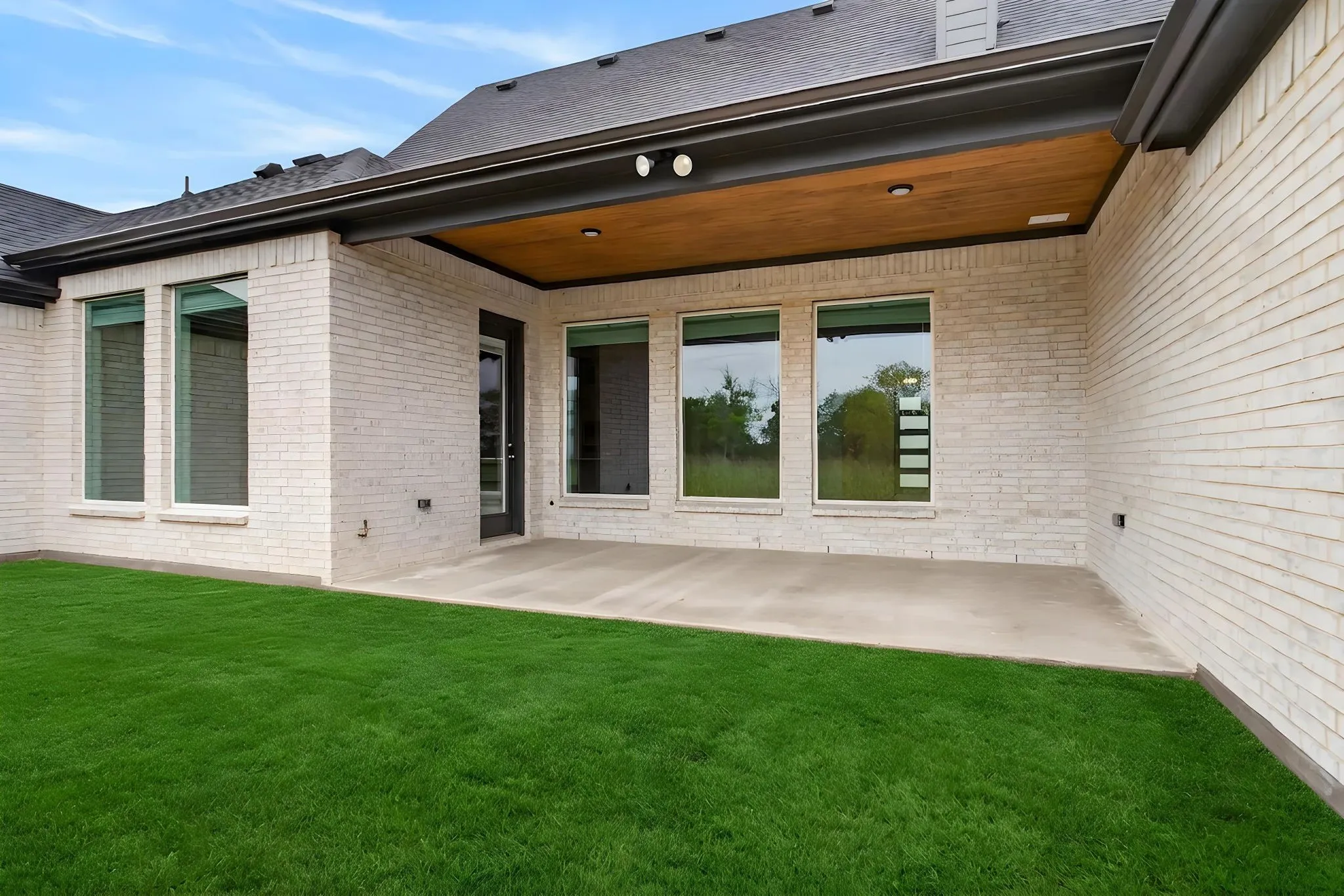 Rear exterior featuring a covered patio with a wood-finish ceiling and recessed lighting, light-colored brick facade, multiple large windows, a glass-paned door, and a manicured lawn