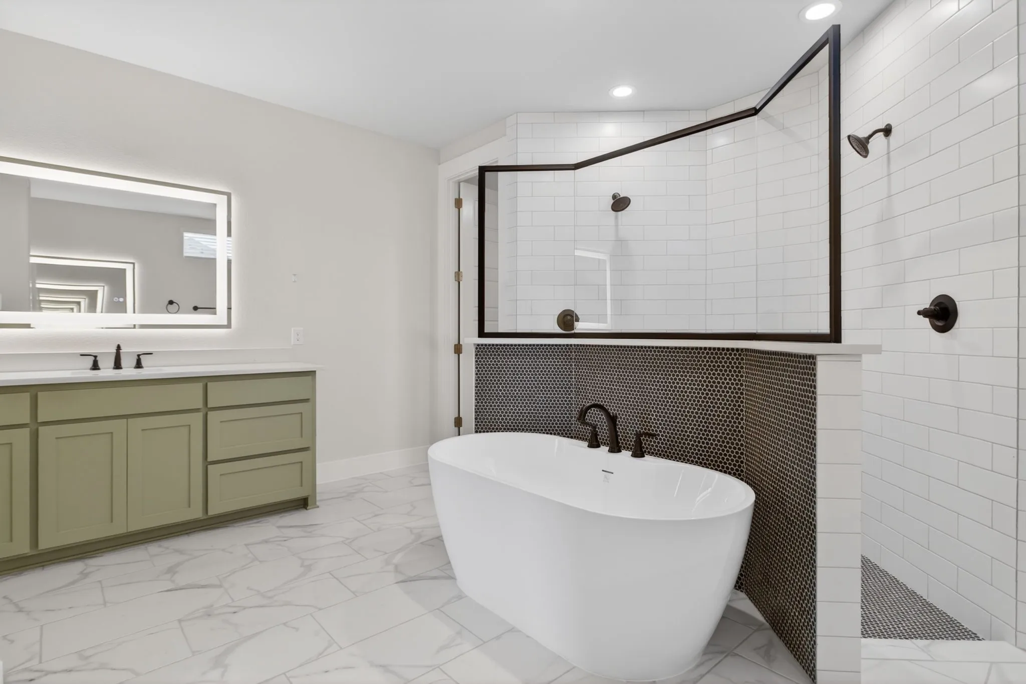 Spacious bathroom featuring a freestanding tub, dual-head shower with subway tile surround, double vanity with a light green finish, back-lit mirror, and patterned floor tile