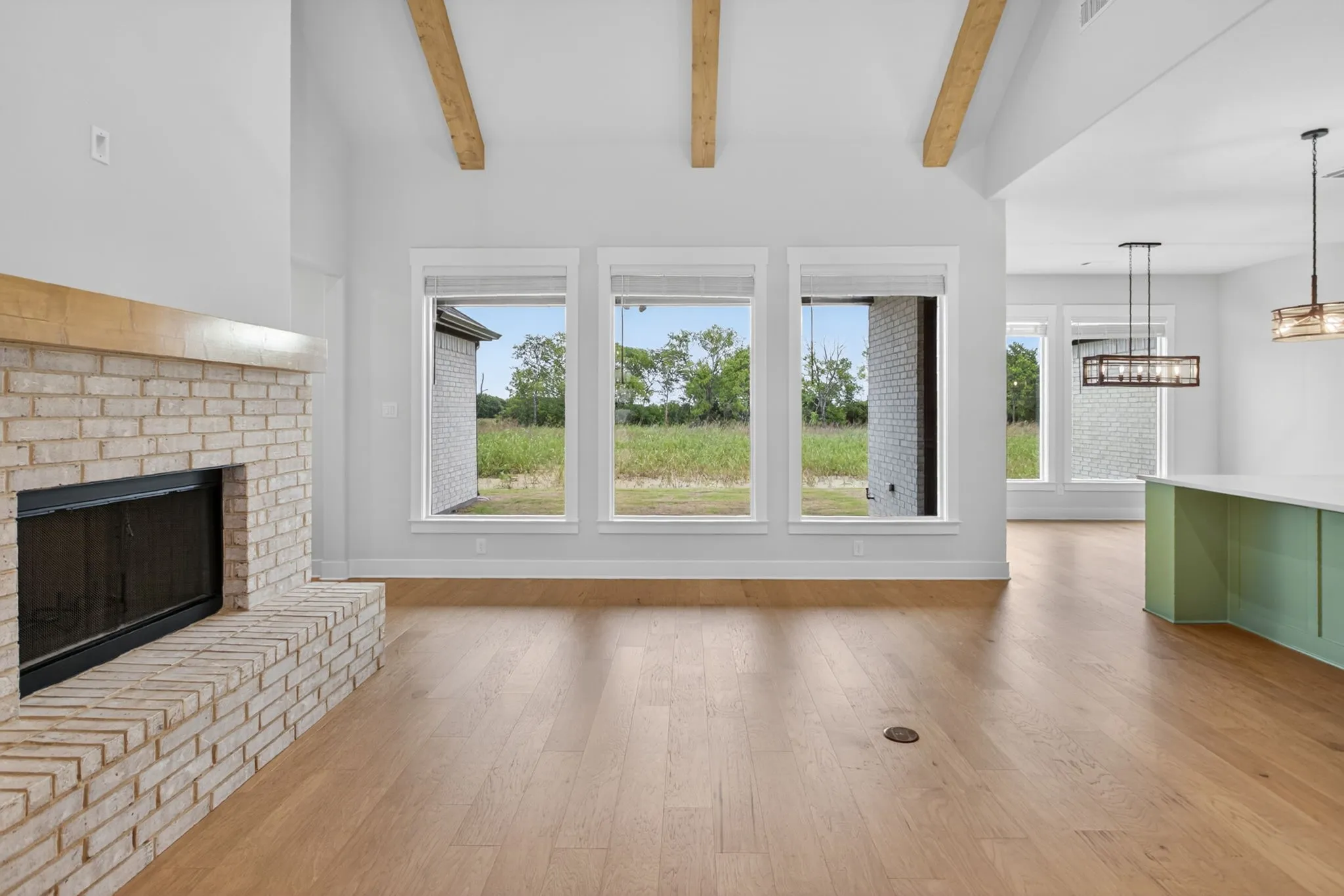 Spacious living area featuring a light-colored brick fireplace with a wood mantel, high vaulted ceilings with exposed wood beams, and light wood-finish flooring