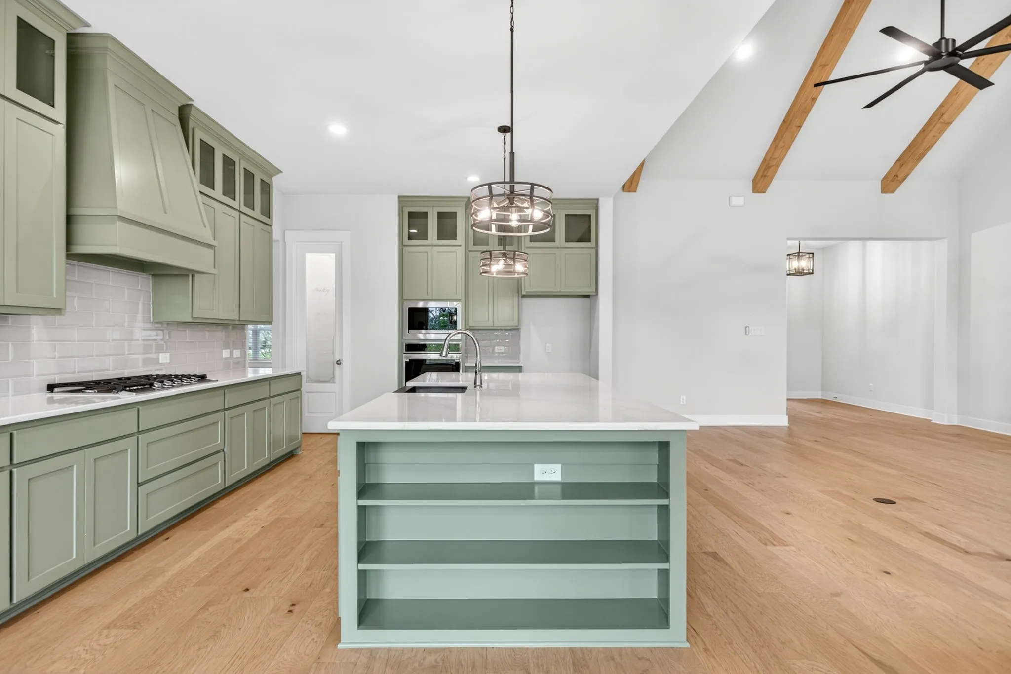 Kitchen and living space featuring a vaulted ceiling with exposed wood beams, a central kitchen island with integrated shelving, light wood-finish flooring, light green cabinetry, and white countertops