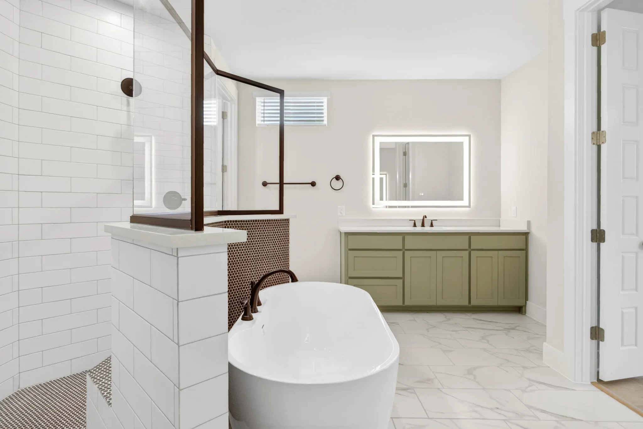 Freestanding soaking tub with dark bronze fixtures, a walk-in shower featuring white subway tile and a dark bronze framed glass enclosure, and a light green vanity with a quartz countertop and an illuminated mirror