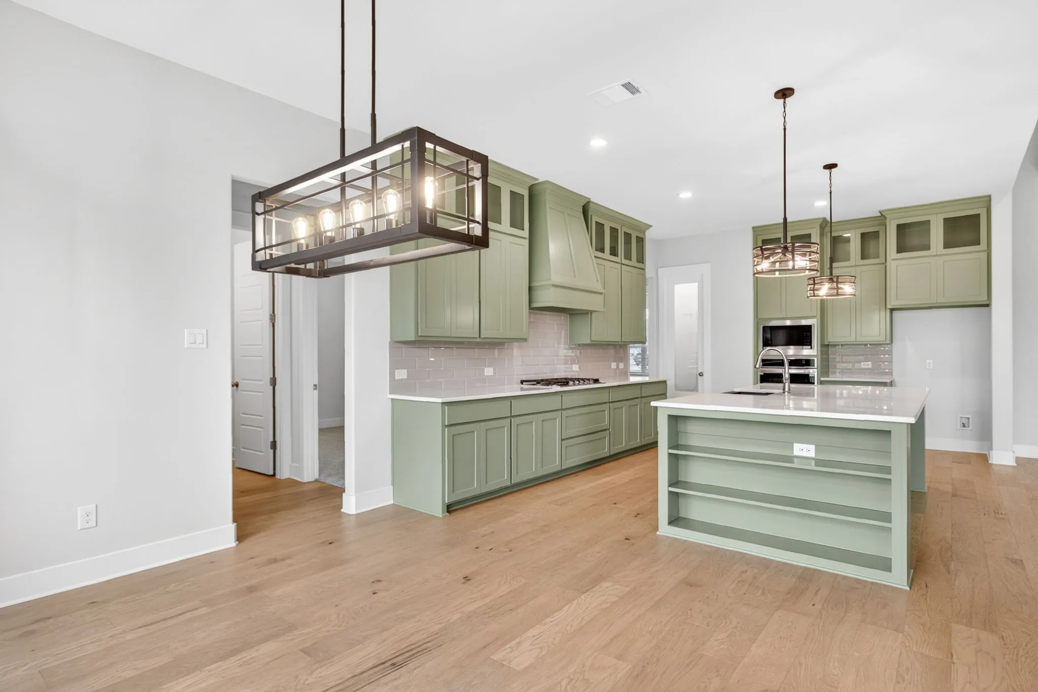 Open-concept kitchen featuring sage green cabinetry, a central island with built-in shelving, light wood-finish flooring, and white countertops