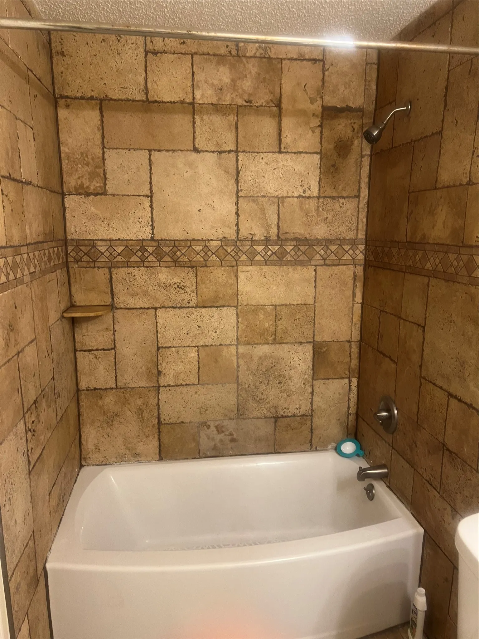 Bathtub and shower combination featuring earth-toned stone-look tile surround with decorative accent band and built-in shower shelf