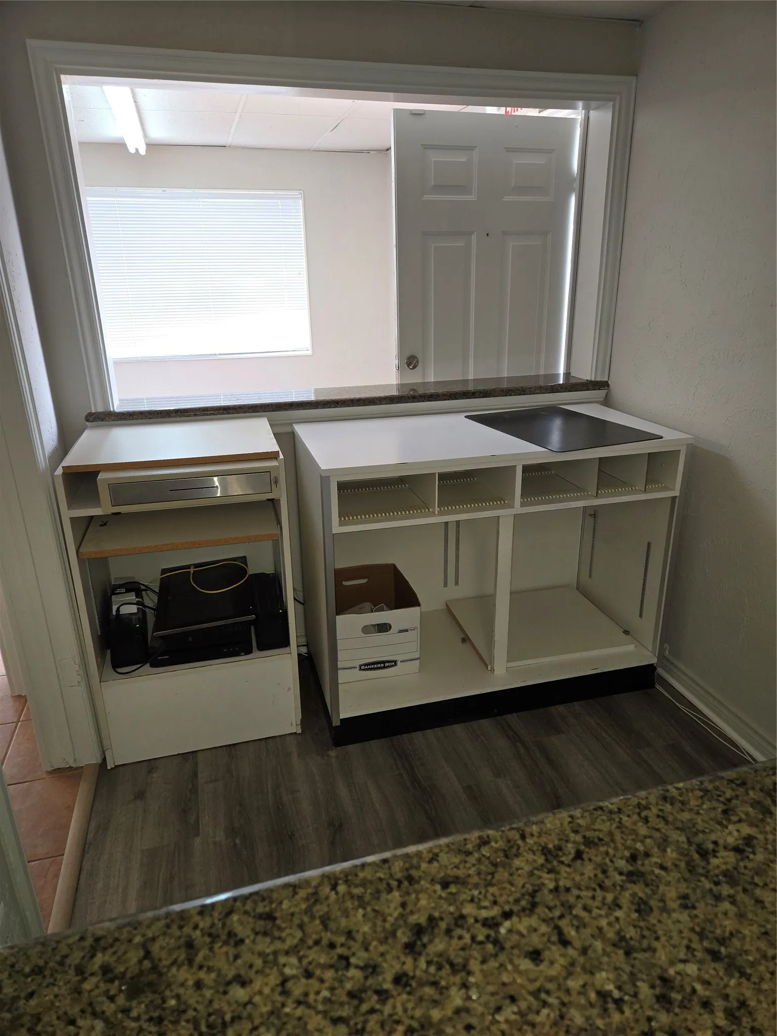 Built-in shelving units under a granite-finish countertop, featuring wood-finish flooring and a pass-through window with horizontal blinds