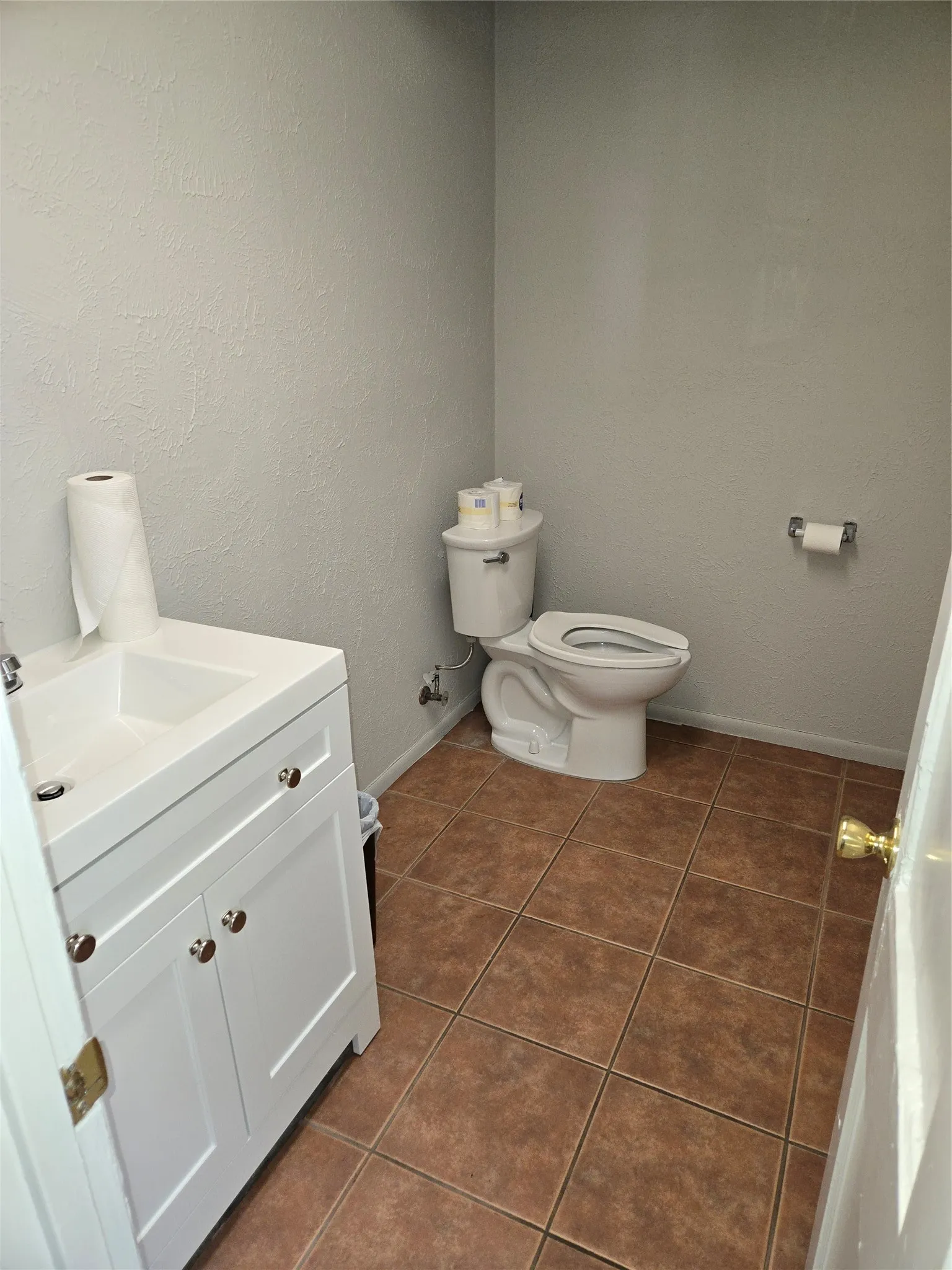 Compact powder room featuring a white vanity with integrated sink, a commode, and brown square floor tiles