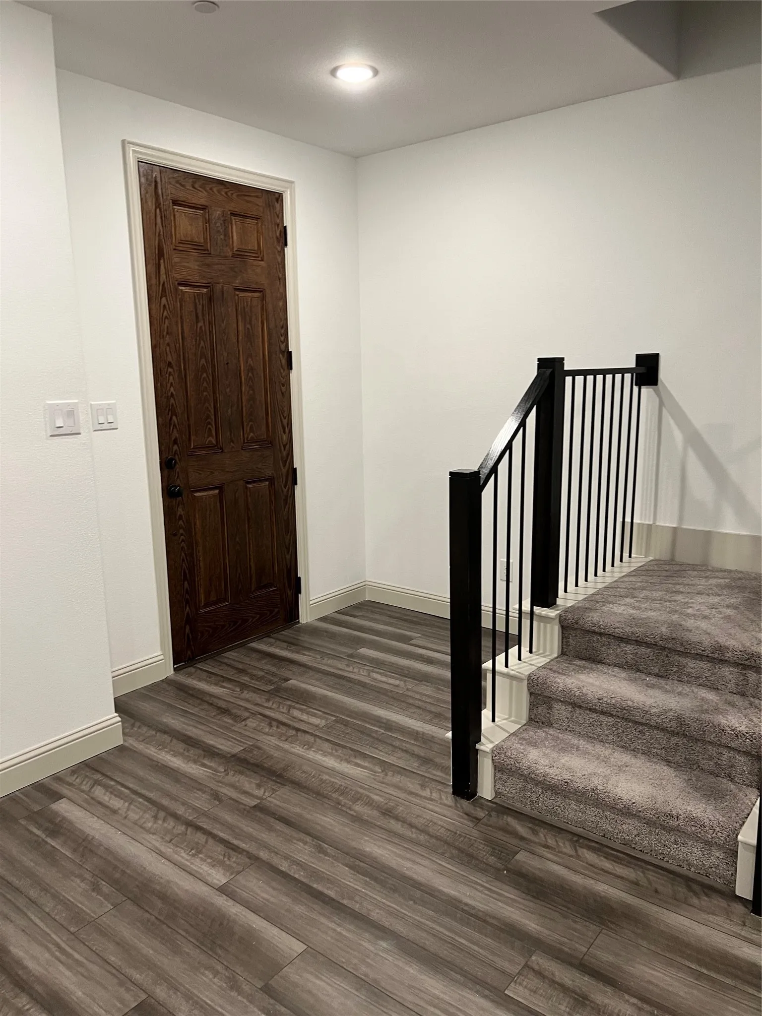Entryway featuring wood-finish flooring, a dark wood paneled door, recessed ceiling lighting, and a carpeted staircase with a black metal railing